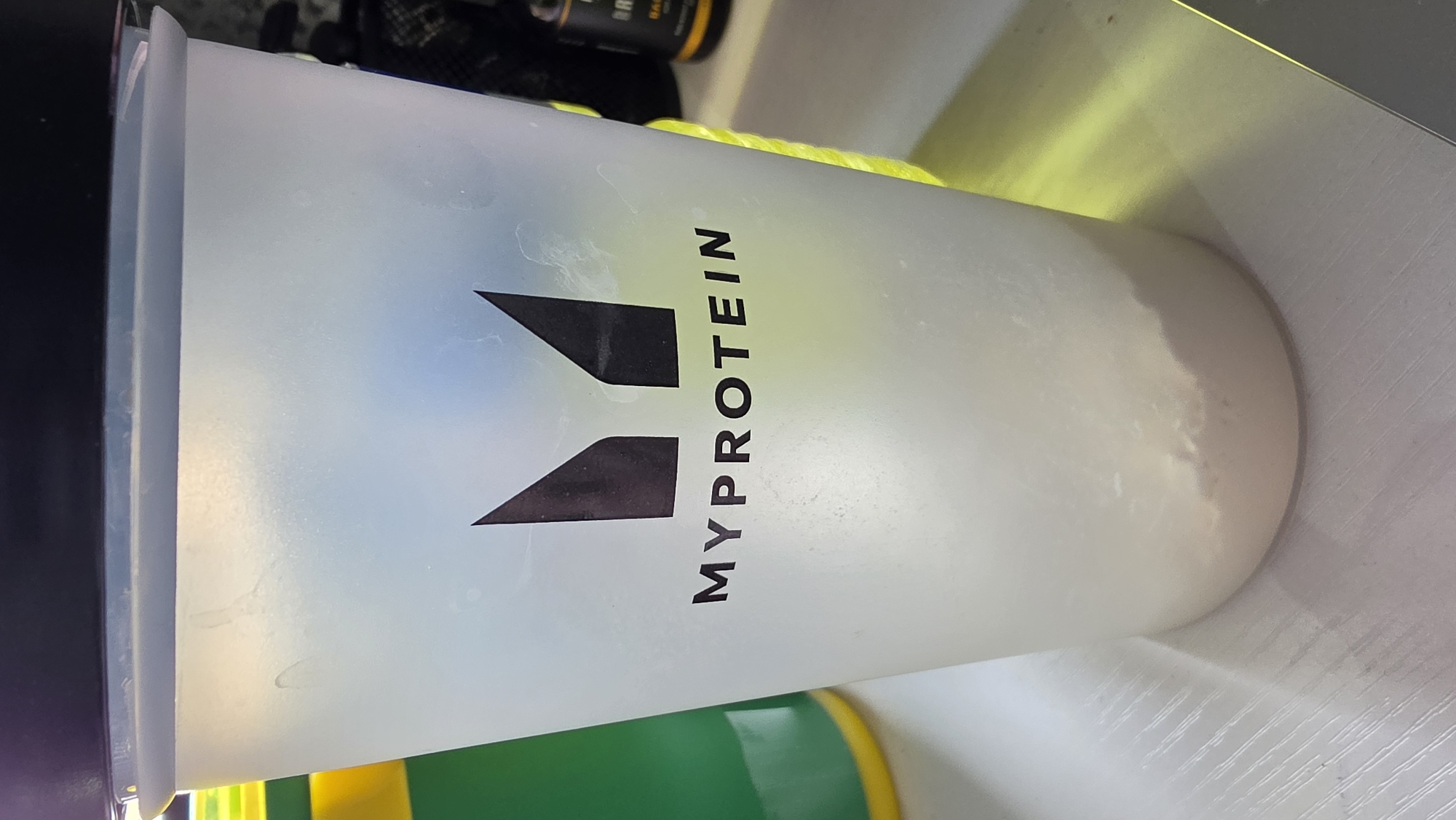The image shows a Myprotein shaker cup on a white surface. The cup is translucent and appears to contain a white powder or liquid. The Myprotein logo is prominently displayed on the side. Other items are vaguely visible in the background, including bottles with labels. The setting appears to be indoors on a flat surface, likely a countertop or shelf. There are no indications of it being located in London or any other specific location. No people are visible.