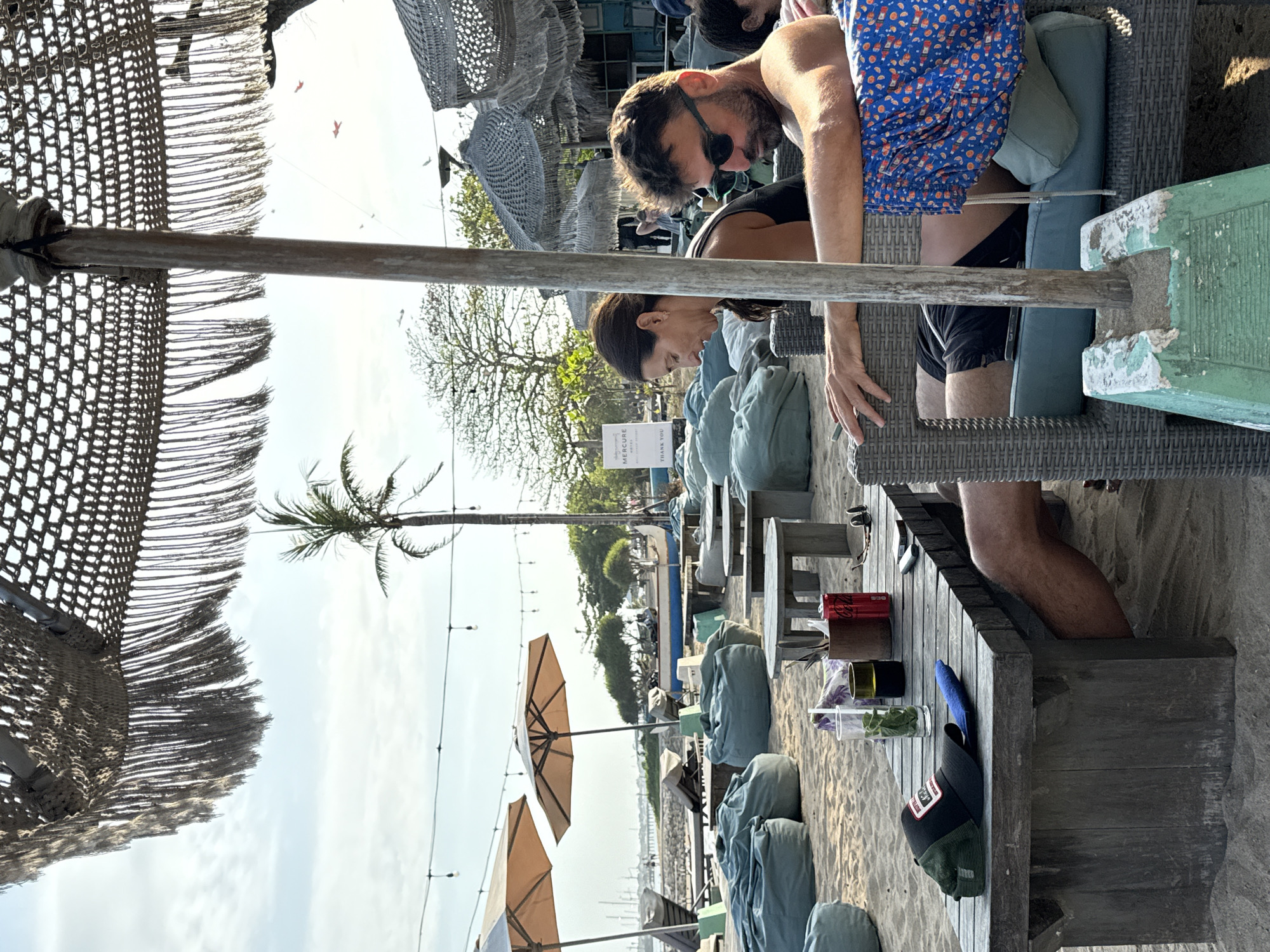A man and a woman are seated at a beachside cafe in Denpasar, Indonesia, under a thatched umbrella. The man, wearing sunglasses and a blue floral patterned shorts, is in the foreground, facing right and looking down. He is seated in a wicker chair with green cushions. The woman, with dark hair, is seated next to him, looking down at a table in front of them. She is wearing a dark tank top. On the wooden table are a glass with mint, a brown cup, a red can of Coca-Cola, a pair of sunglasses, and a black baseball cap. In the background, there are more beach chairs, beanbag chairs, and tables on the sand. Palm trees and other foliage are visible, as is a blue and white boat. The sky is bright and cloudy.