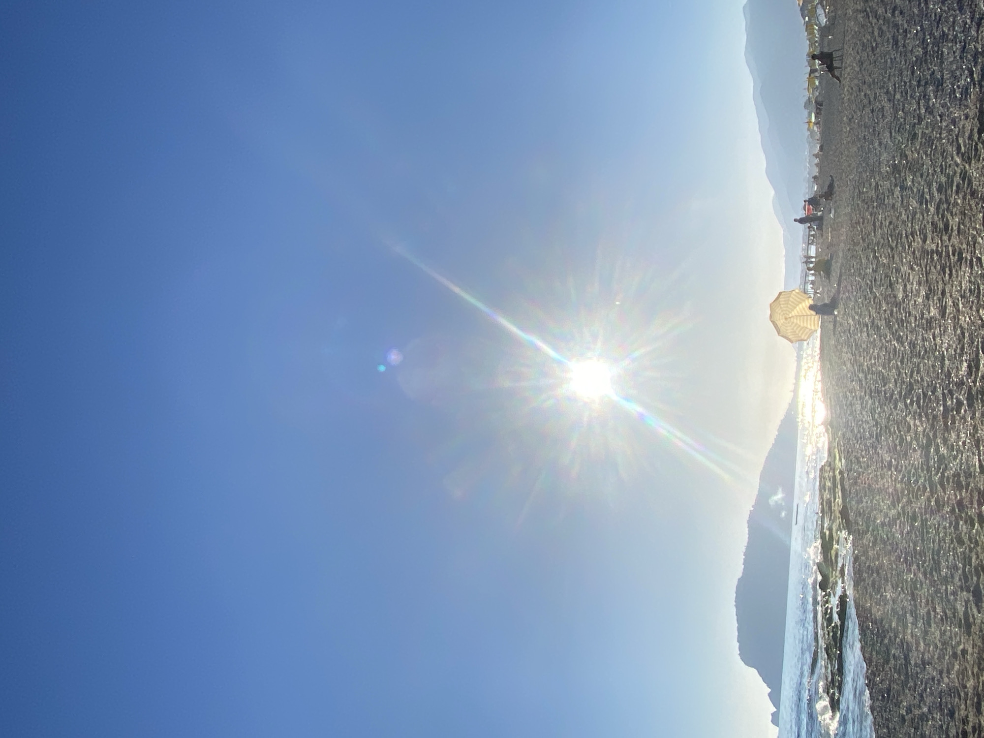 The image depicts a beach scene in Alanya, Turkey, taken on a sunny day with the sun prominently in the frame. The sky is a clear blue. To the right is the beach, composed of dark sand and water. Mountains are visible in the background. Along the beach, there are figures of people, and beach umbrellas are scattered across the sand. There are waves near the shore.