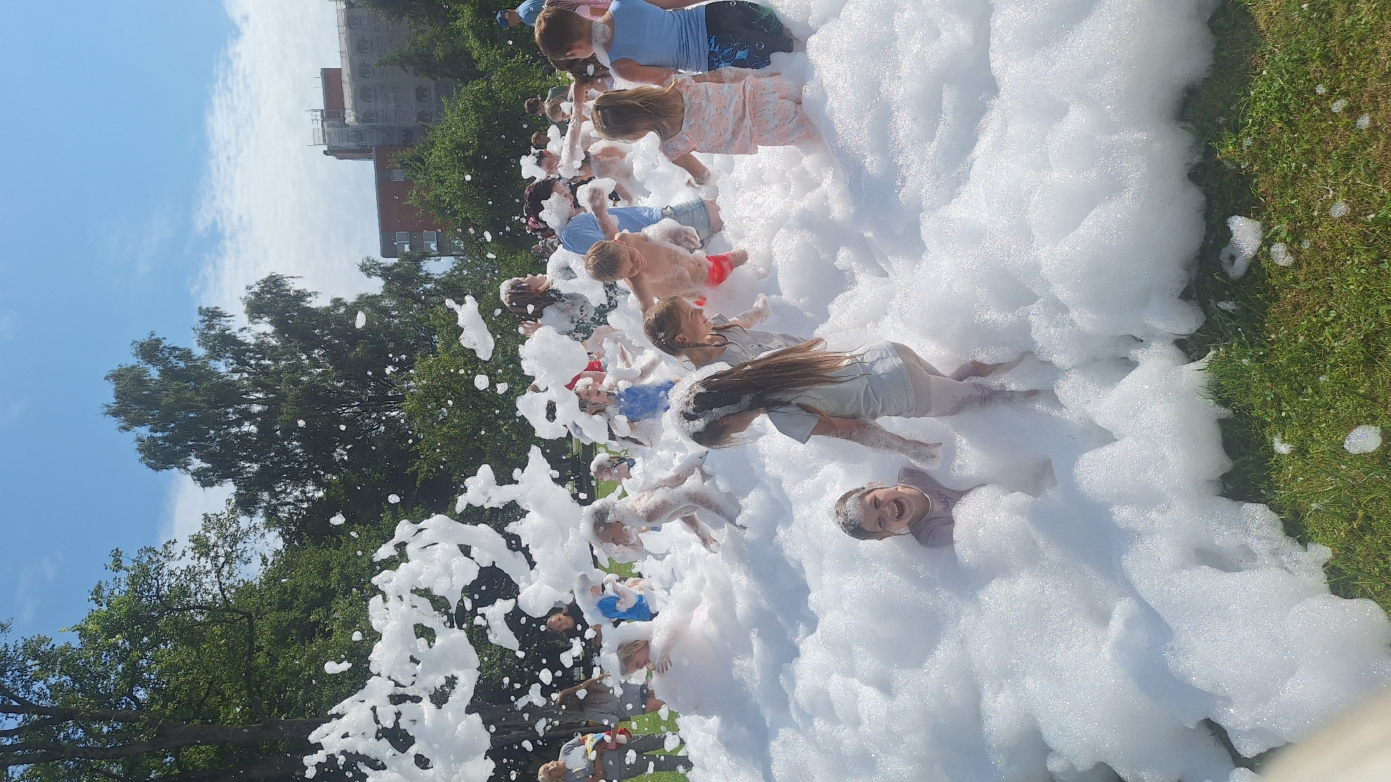 In Lēdmane, Latvia, on a sunny day with a partly cloudy sky, a group of children are joyfully playing in a large mound of white foam. The foam covers most of the lower half of the image, creating a playful, immersive environment. Children of various ages are visible, some partially submerged in the foam, others standing or moving through it. They are dressed in casual summer clothing, including swimsuits and t-shirts. The children are interacting with each other and the foam, with smiles and laughter evident on many faces, indicating a fun outdoor activity, likely a foam party. In the background, trees and some buildings are visible, suggesting an urban or suburban park setting. The ground beneath the foam appears to be grass.