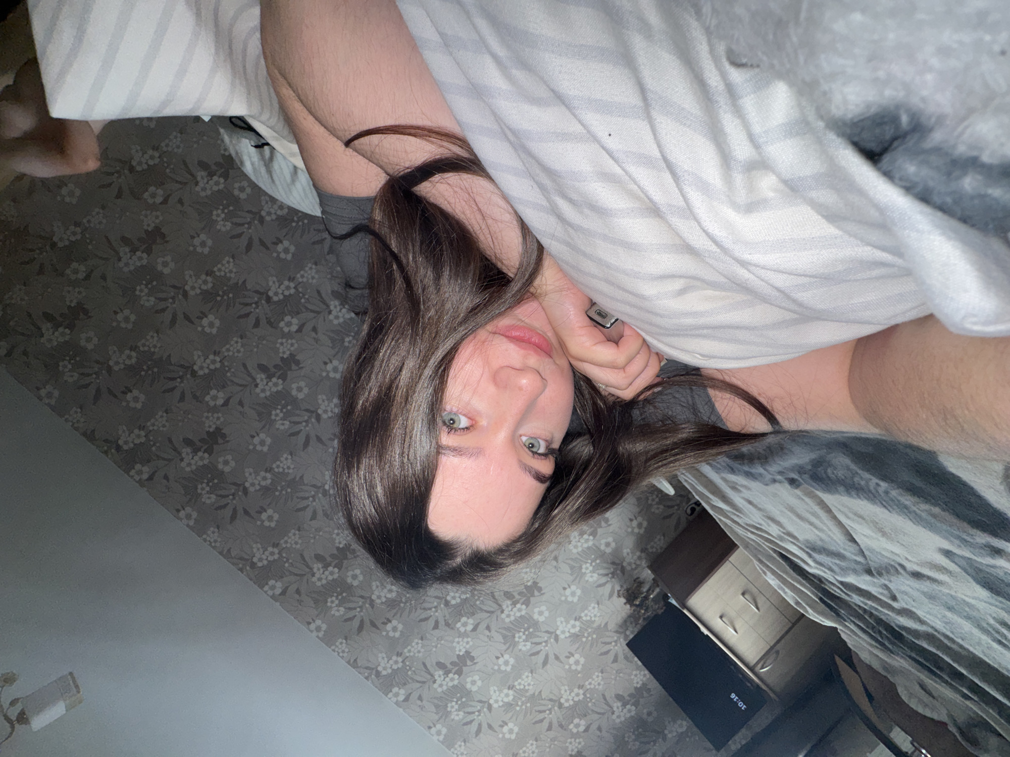 The image is a selfie taken indoors, likely in a bedroom in Башкултаево, Russia. A young woman with long, dark brown hair and light blue eyes is lying on a bed, propped up on her arm. She is wearing a gray t-shirt. Her head is tilted, and she is looking towards the camera with a slight smile. The background shows a wall with floral wallpaper and a glimpse of a dresser and a television. The bed has striped pillows and a gray blanket. The overall lighting suggests it might be evening or a dimly lit room.