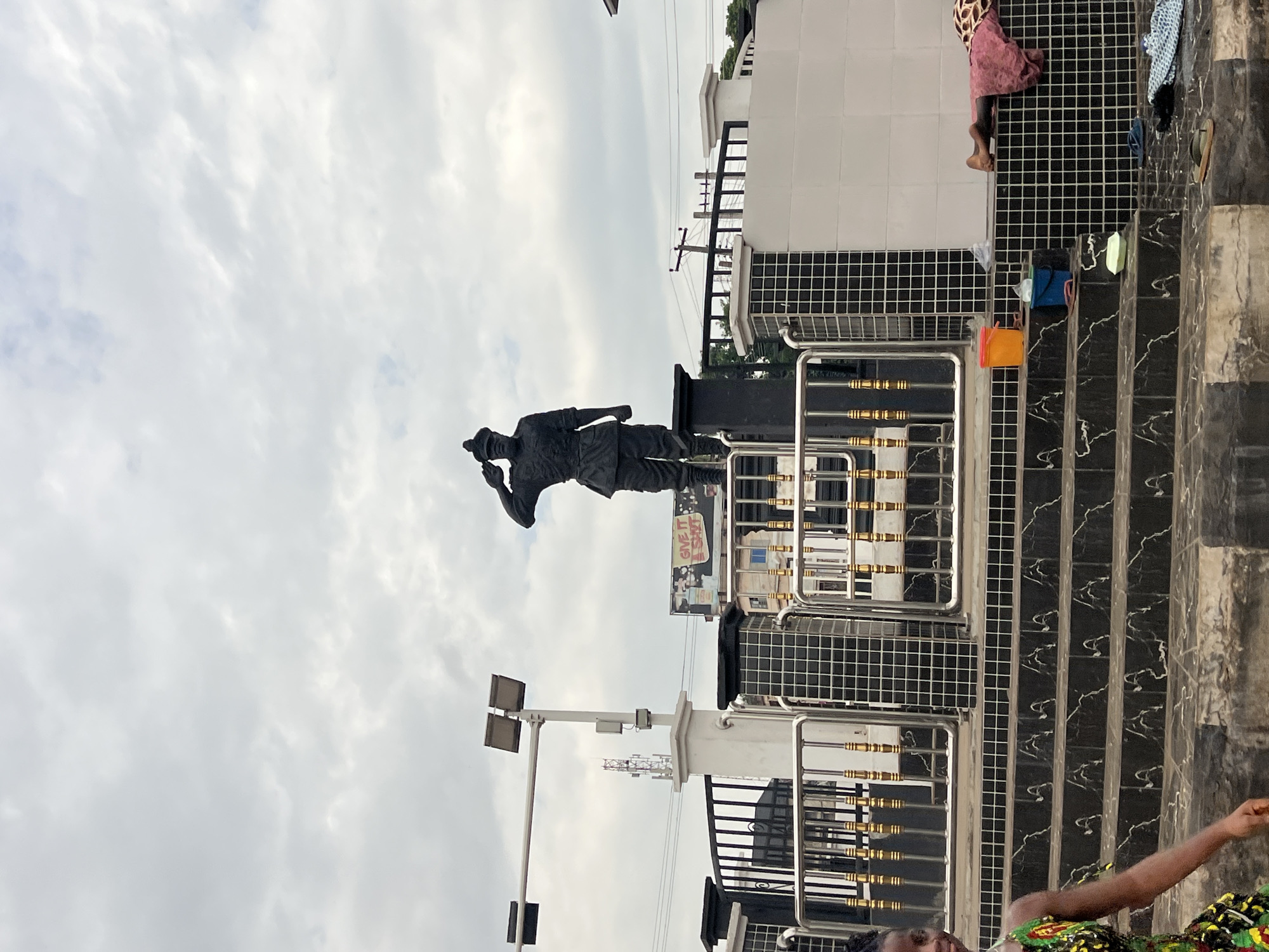 A statue of a military figure saluting stands atop a tiled platform with metal railings in Nkpor, Nigeria. The statue is dark and silhouetted against a cloudy sky. In front of the statue, there are steps with black and white patterned tiles. To the left of the steps, a young woman in a colorful dress walks by. To the right of the steps, there are two buckets, one orange and one blue, and a person is lying down on a tiled surface, partially covered by a pink and patterned cloth. Several pairs of footwear are scattered on the ground. In the background, a traffic light and utility poles are visible, along with buildings and a billboard that reads "GIVE IT A SHOT".