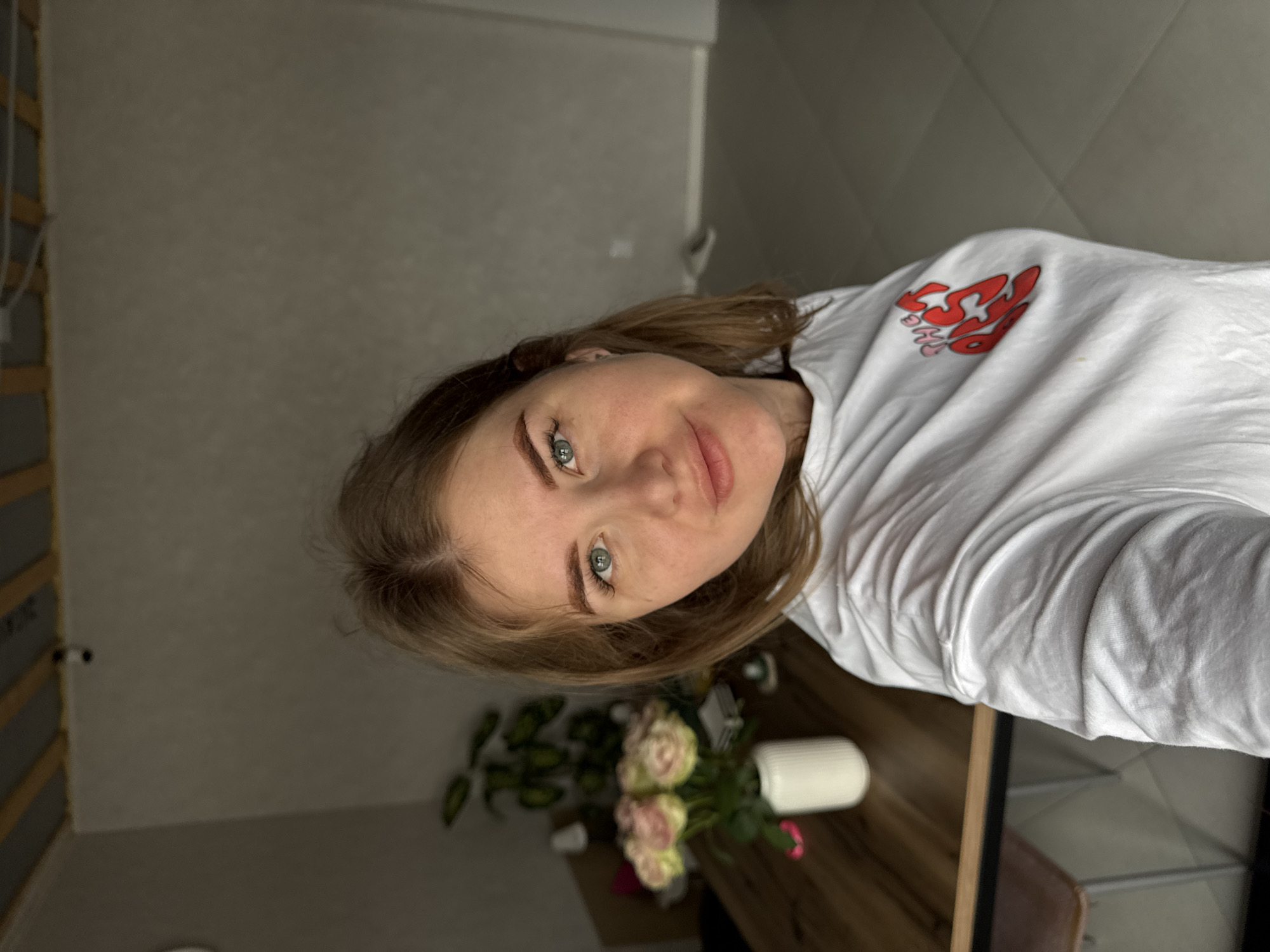 A woman with fair skin, light brown hair, and green eyes is centered in the image, wearing a white t-shirt with a red design on the upper right. She is indoors, looking towards the camera with a slight smile. Behind her, there's a wooden table with a vase of pink and white roses. The background includes a grey wall and a section of what appears to be a door or wardrobe with a light wood frame. The location is Gornyi (Dvurechenskoe S/P), Russia.