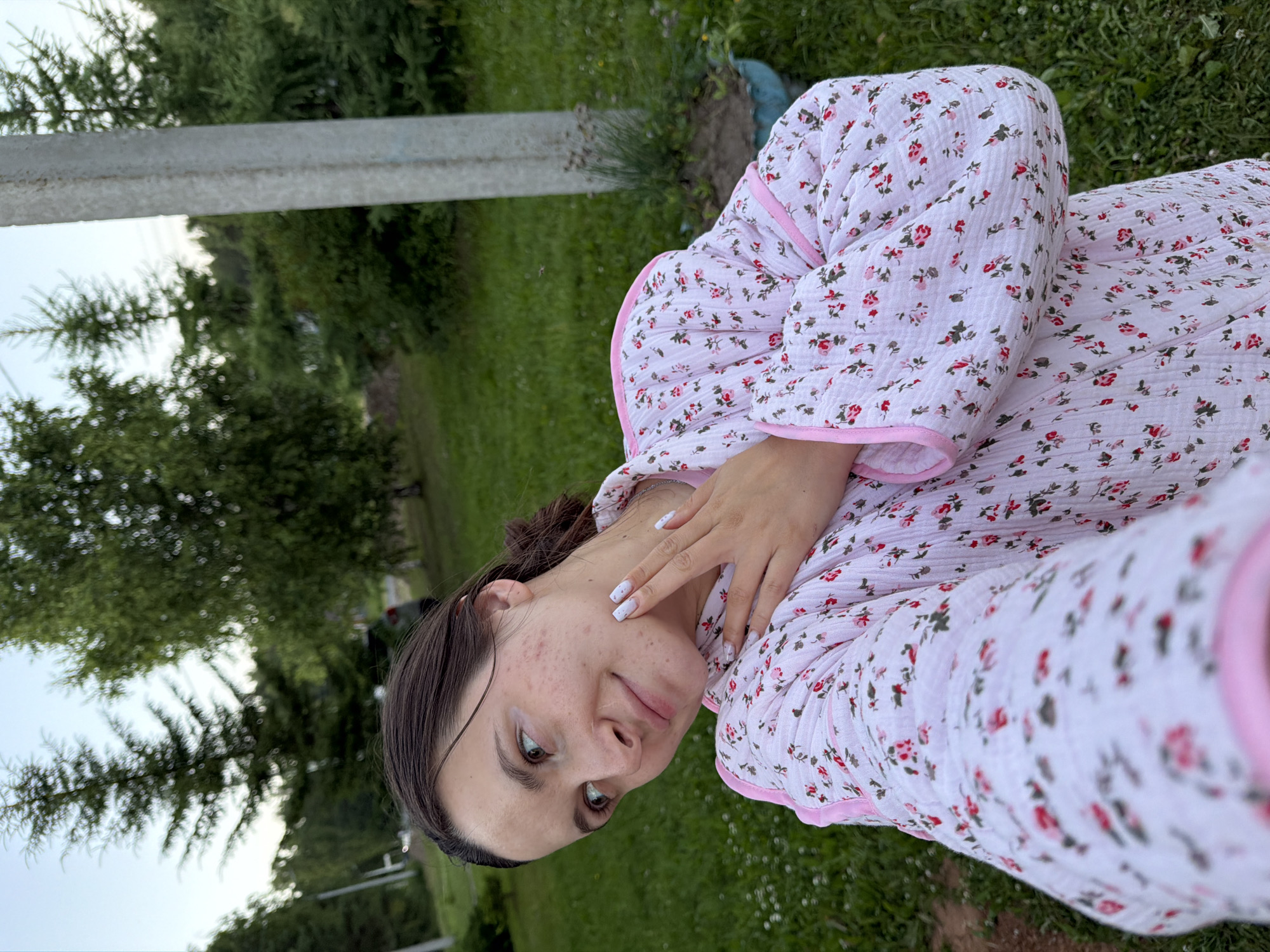 A young woman with fair skin and brown hair pulled back in a bun is captured in a close-up, outdoor selfie. She is wearing a white quilted robe with a pink floral pattern and pink trim. Her left hand is raised, touching her neck, and her fingernails are painted white with small black speckles. She is looking to her left, with her head tilted slightly. The background consists of a grassy area with scattered small white flowers, tall green trees, and a light gray pole on the right. The lighting suggests it is either early morning or late afternoon. The scene appears to be a residential area within the city of Krasny Mayak, Russia, possibly a backyard or a park given the vegetation.
