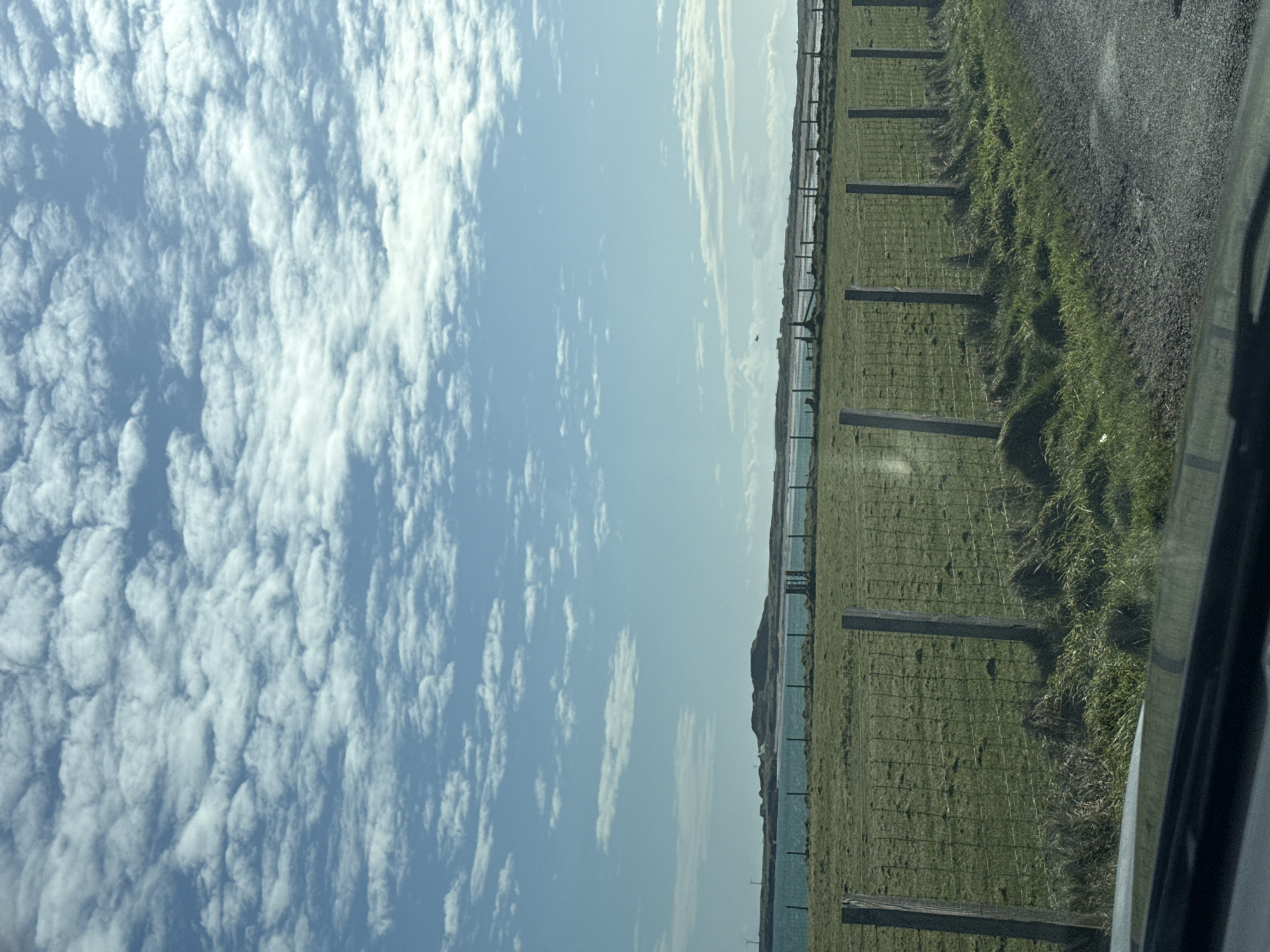 The image shows a sky with a dense blanket of altocumulus clouds covering the left side of the frame, transitioning to a clearer, light blue sky toward the right. At the right, a strip of land is visible, featuring a grassy area delineated by a fence with wooden posts. This grassy area seems to border a body of water, possibly a sea or ocean. A road runs alongside the grassy area, and part of a car's interior is seen in the right foreground, suggesting the image was taken from within a vehicle. The location is Unknown, Unknown.