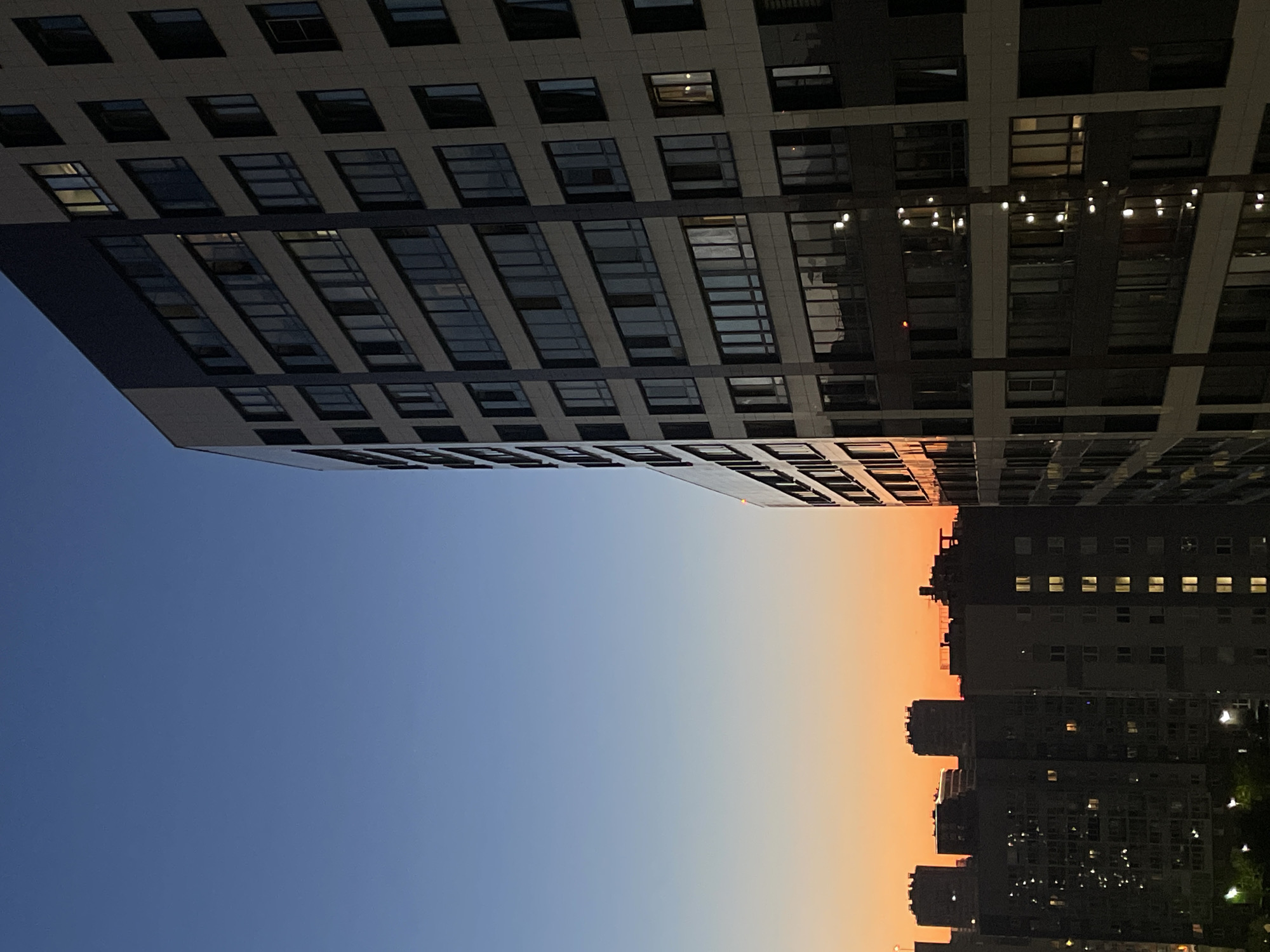 The image captures an urban landscape at dusk, featuring a series of multi-story buildings against a gradient sky that transitions from deep blue at the top to a warm orange near the horizon. In the foreground and to the right, a modern high-rise building with a beige and dark grey facade dominates the frame. Its numerous windows, many of which are lit, reflect the ambient light. To the left and in the background, other buildings of varying heights and architectural styles are visible, silhouetted against the fading light. Some of these buildings also have illuminated windows, suggesting residential occupancy. The scene appears to be a residential area within the city of Sinyavino, Russia, characterized by dense urban development and the transition from day to night. There are no visible people or overt actions taking place.