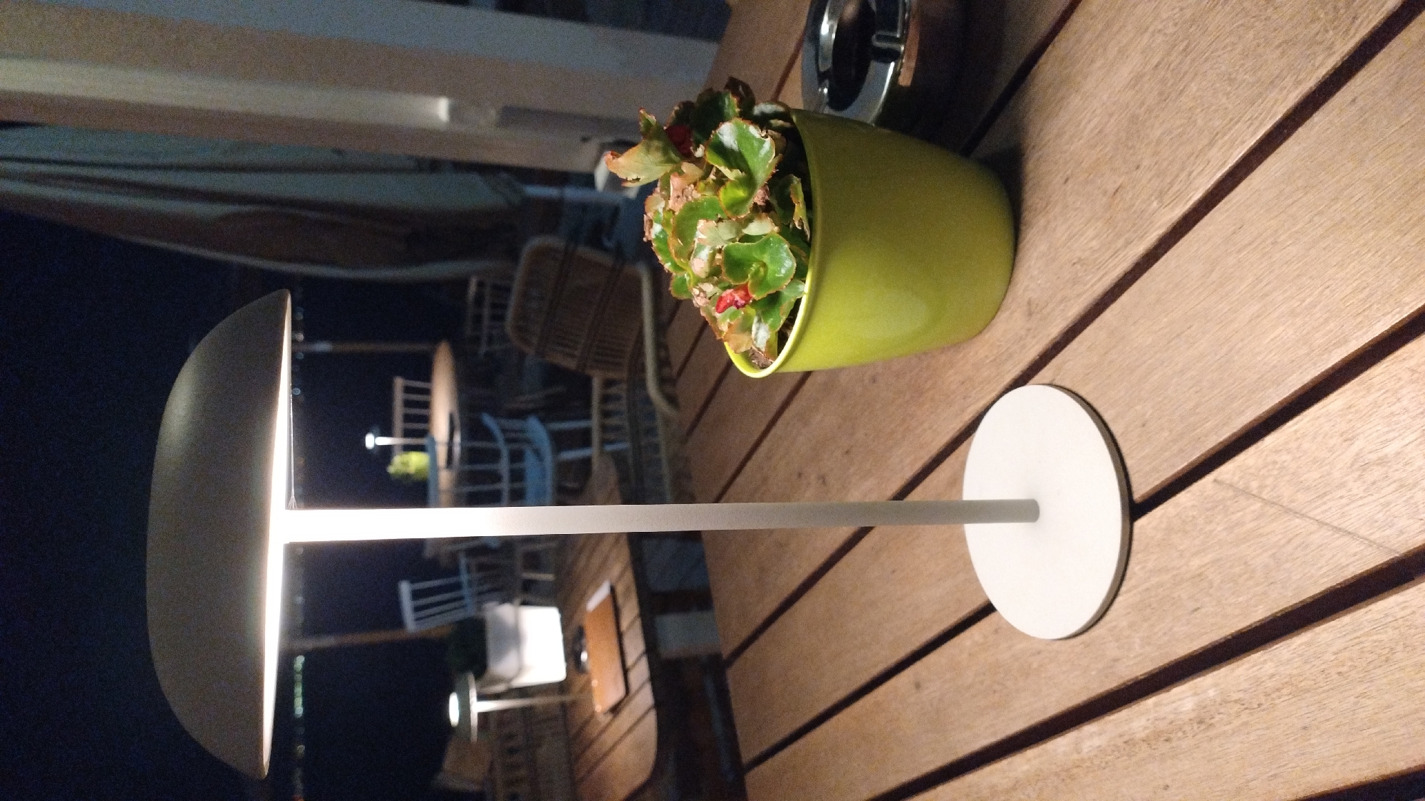 A close-up, eye-level shot captures a wooden outdoor table at night. A modern, dome-shaped, white lamp with an illuminated top stands centrally on the table, casting a soft glow. To the right of the lamp, a bright lime green pot holds a lush, leafy green plant with small red flowers. An ashtray is visible to the right of the plant. In the background, out-of-focus tables and chairs suggest a restaurant or cafe setting, possibly with umbrellas for shade. The dark sky indicates it is nighttime. This scene is situated at a restaurant in Perea, Greece.
