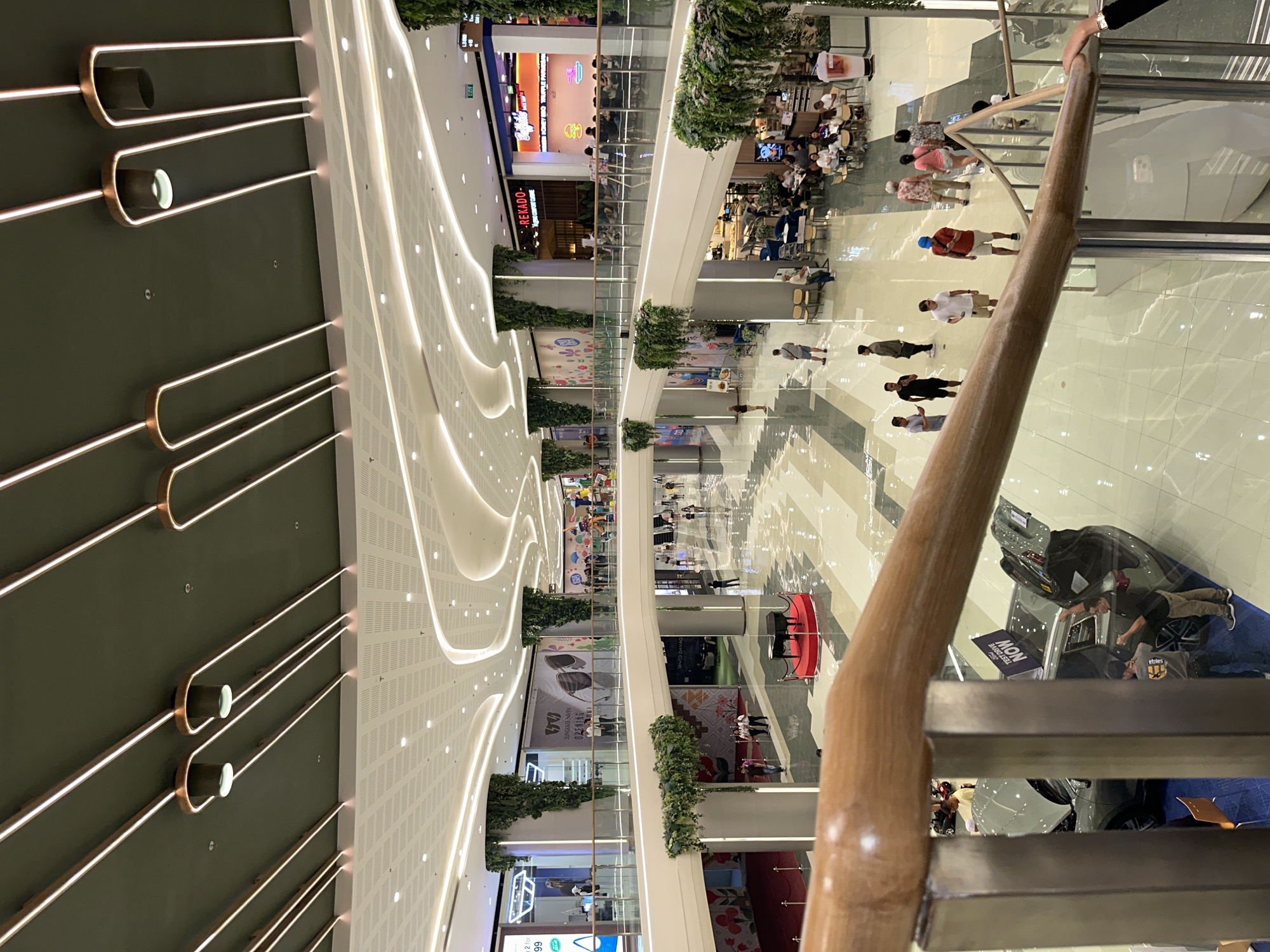 This is a photograph taken inside a modern, multi-level shopping mall, likely in Davao City, Philippines, given the presence of "DAVAO CITY" text visible on a banner. The image captures a wide interior view, showcasing the mall's architecture with a dark green ceiling adorned with decorative, illuminated light fixtures. The lighting is bright and modern, with white linear lights along the ceiling and ambient lighting throughout the mall.
The mall features several levels connected by escalators and walkways. Lush green vertical gardens are incorporated into the design, with plants cascading down large cylindrical columns that support the upper levels. The floors are polished and reflective, contributing to the bright and spacious feel of the environment.
Several people are visible throughout the mall, engaged in various activities such as walking, shopping, and dining. On the lower level, a display featuring a car with its trunk open is set up, with a banner indicating "TEST DRIVE NOW." A black grand piano on a red circular platform is also visible in the central area of the lower level.
In the foreground, a wooden handrail of a staircase is prominent, suggesting the photograph was taken from an elevated position looking down into the mall. A person's hand with a black bracelet is visible on the right side of the frame, near the handrail. The overall impression is of a bustling and well-maintained commercial space.