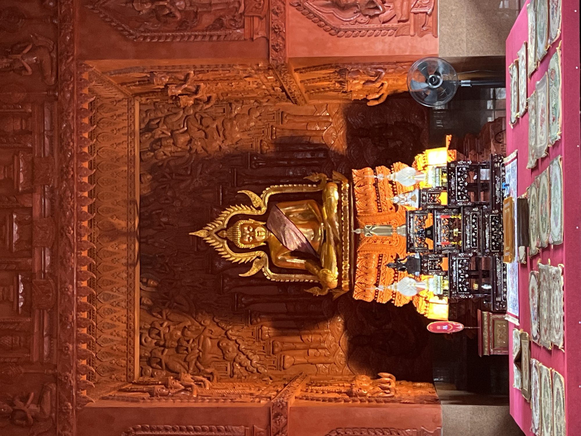 The image shows a golden Buddha statue seated on a pedestal in an ornate, dimly lit chamber. The chamber is decorated with intricate wood carvings depicting figures and scenes. In front of the Buddha statue is an altar with several objects, including smaller statues, candles, and what appear to be religious texts laid out on a red-draped table. To the right of the altar, a black oscillating fan is visible. The overall color palette is dominated by warm, reddish-orange tones, likely from the wood and lighting. The scene appears to be inside a temple or shrine in Baan Hua Thanon, Thailand.