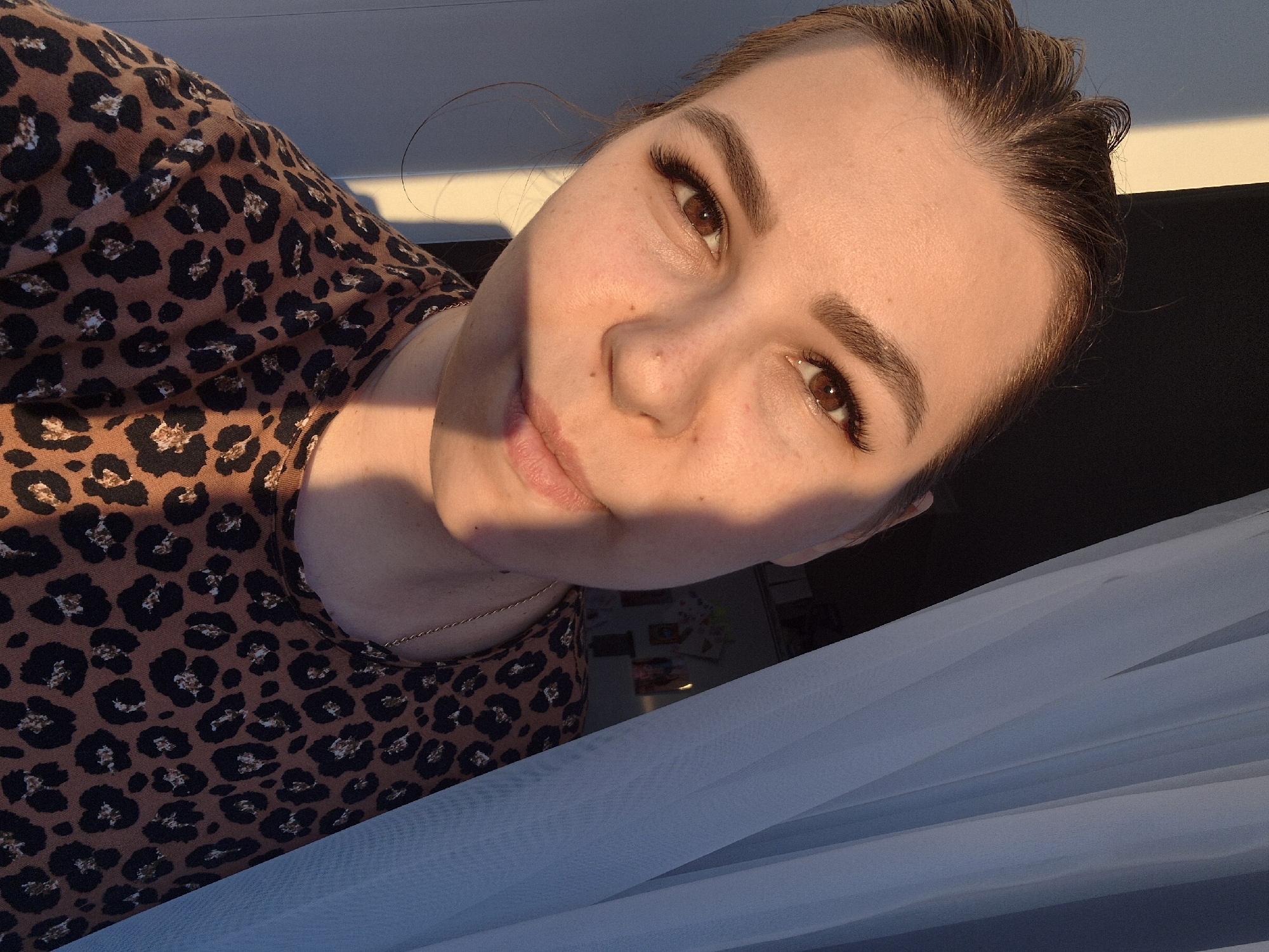 A person with fair skin, brown eyes, and light brown hair pulled back into a bun is facing the camera. The person is wearing a tan shirt with a black leopard print pattern. A thin gold chain necklace is visible. Sunlight is casting shadows across the face and shirt. Part of a white or light blue curtain is visible at the bottom right of the frame. No specific location in Tobolsk, Russia is discernable from the image.