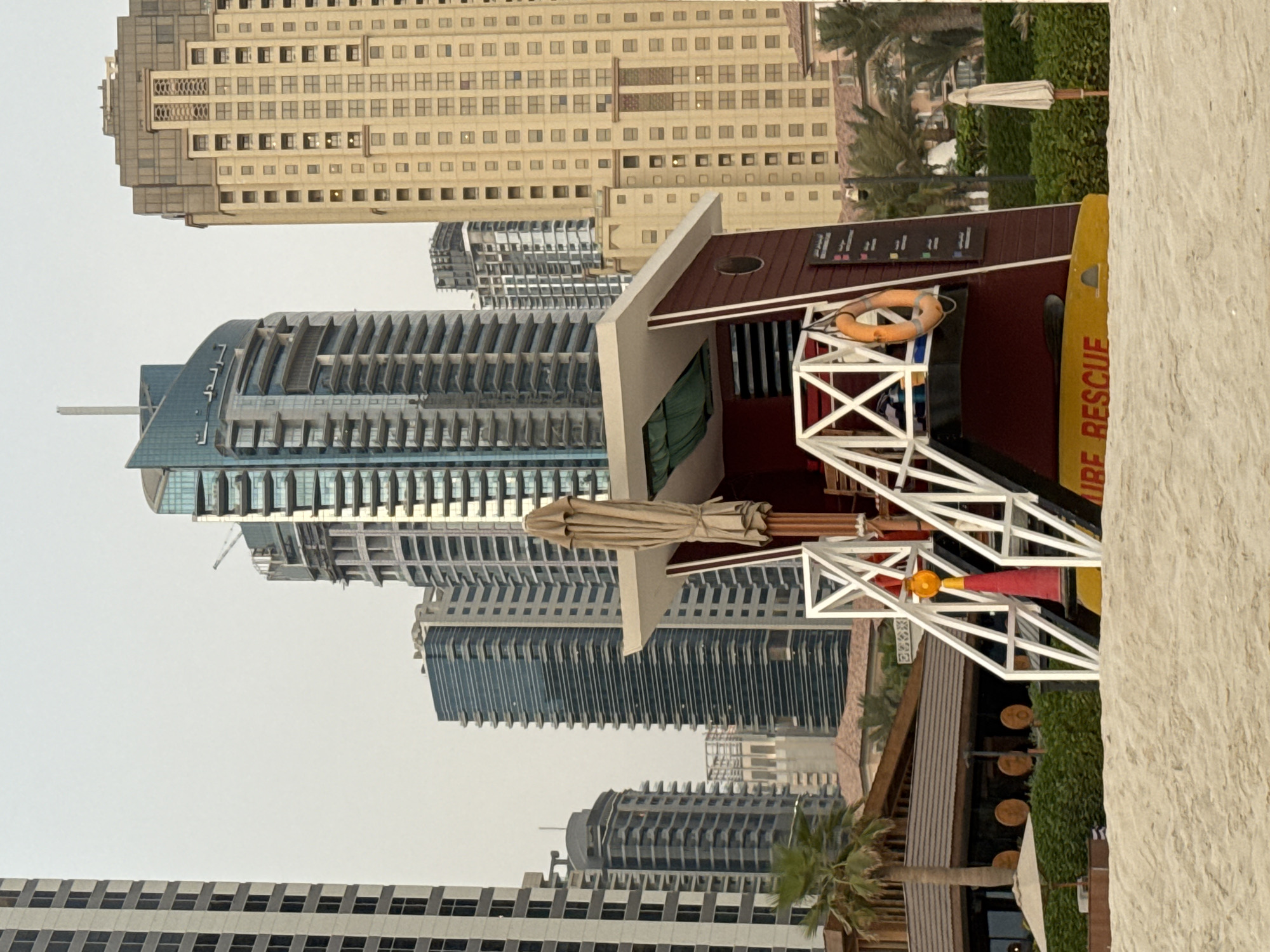 A lifeguard tower stands on a sandy beach in Dubai, United Arab Emirates, with towering skyscrapers in the background. The tower is painted maroon with white railings and a yellow base displaying the words "LIFE RESCUE" in red. An orange life preserver hangs on the side of the tower. A closed umbrella rests on the tower's roof. In the distance, several modern high-rise buildings, characteristic of Dubai's skyline, are visible, including a distinctive building with a curved, glass facade. Palm trees and lush green bushes line the area between the beach and the buildings. The sky is overcast, suggesting it may be early morning or late afternoon.