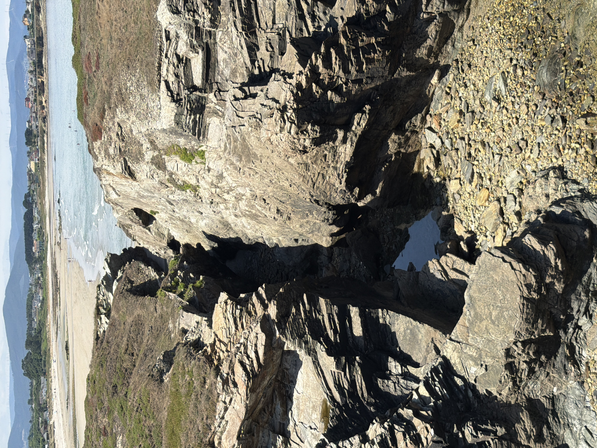 This is a high-angle, eye-level, medium shot of a rocky cove leading to a beach with a town in the background. The rocky cove is a prominent feature in the foreground, with layered rocks forming a dramatic, deep chasm. A small pool of water is visible at the bottom of the chasm. The rocks are a mix of grey and brown tones, with some green vegetation growing on the upper slopes. The beach is sandy with gentle waves lapping at the shore. Several people are visible on the beach and in the water. In the background, a coastal town with houses and buildings is nestled against rolling hills. The sky is clear and blue, with some clouds visible. The scene is likely located on the coast of Ribadeo, Spain, known for its dramatic rock formations and beaches.