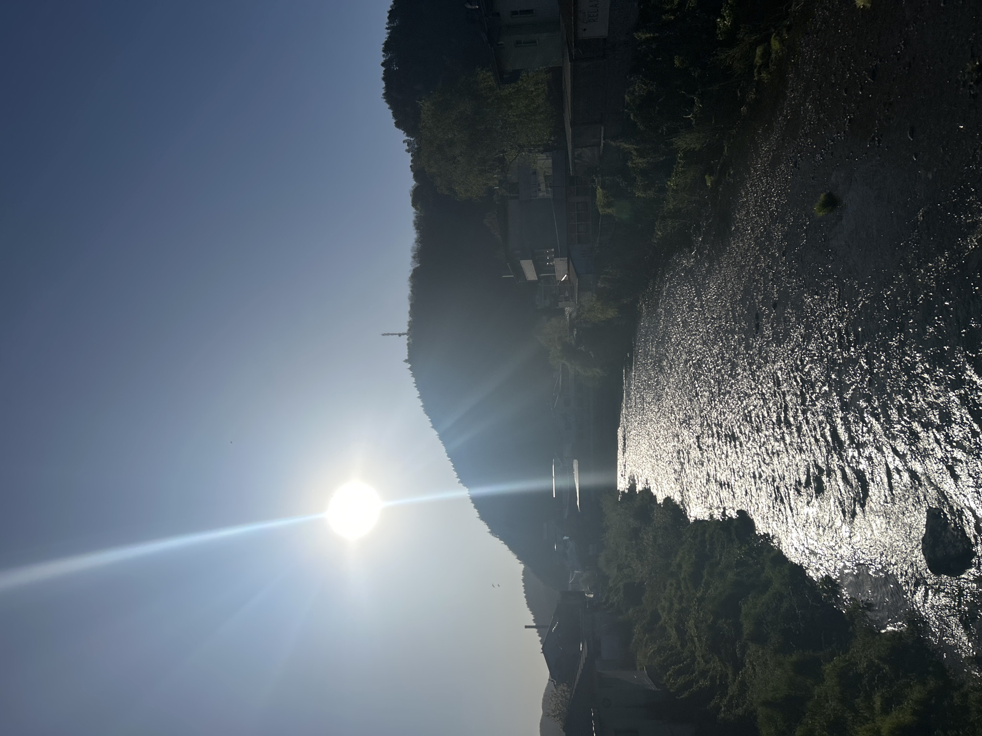 The image shows a river in Zlatograd, Bulgaria, glistening under the bright sunlight. The sun is low in the sky, casting a long glare across the water. Trees line the riverbank, with buildings visible on the hillside.