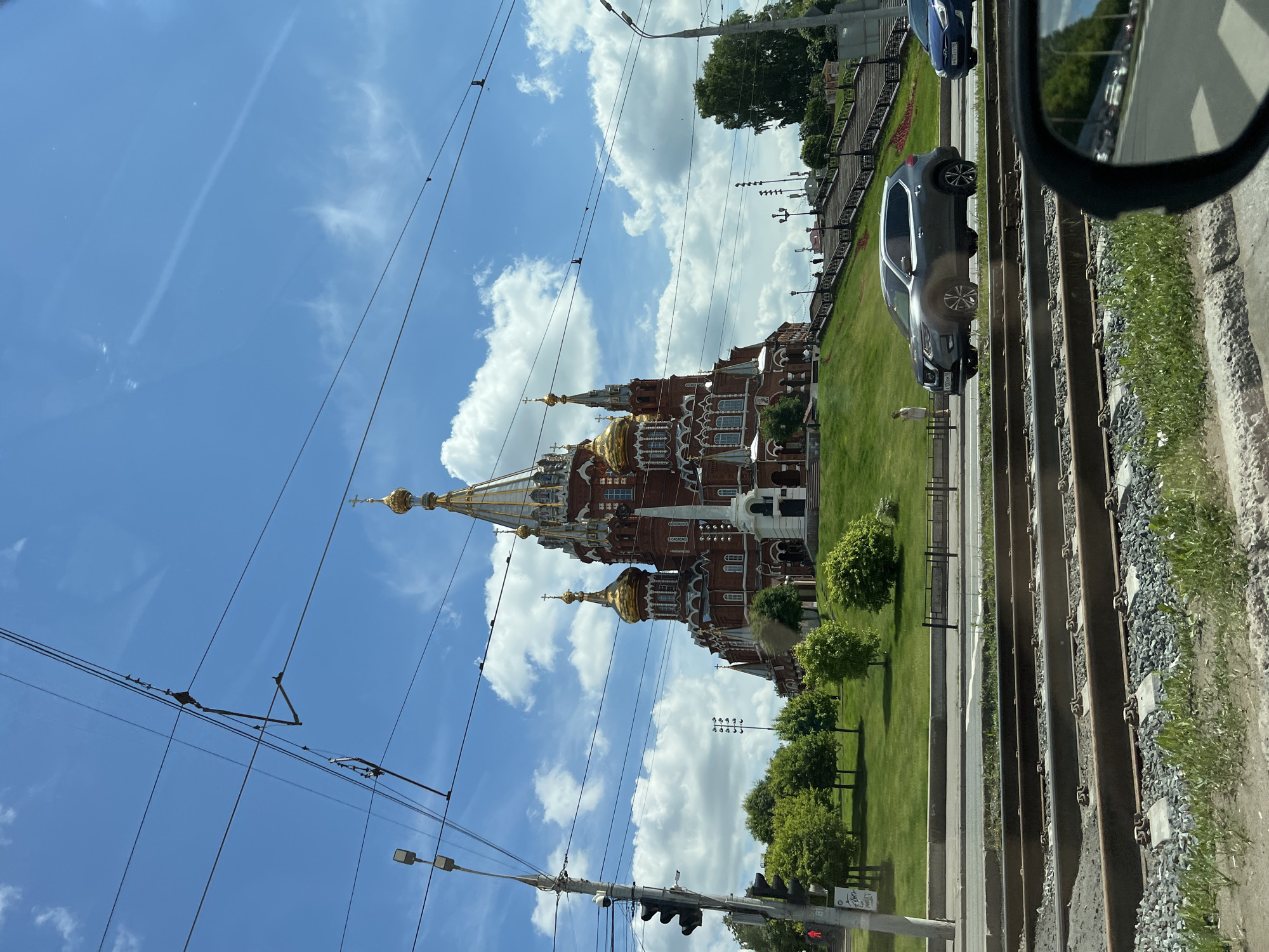 The image displays a bright, sunlit urban scene in Izhevsk, Russia. A magnificent red-brick cathedral, likely St. Michael's Cathedral, with numerous golden domes and spires, stands prominently on the left side, surrounded by a vibrant green lawn and scattered trees. The cathedral's ornate architecture suggests a Russian Orthodox style.
In the foreground, a set of tram tracks runs horizontally, indicating a railway or tram line. Adjacent to the tracks is a paved road where several vehicles are visible. A dark grey SUV, possibly a Nissan, is centrally positioned on the road, with its license plate partially readable as "A 120 OP 18", signifying the Udmurt Republic where Izhevsk is located. Another blue car is partially visible further down the road. The photograph appears to be taken from inside a vehicle, as a car's side mirror occupies the bottom right corner of the frame.
The sky is a clear, brilliant blue with scattered fluffy white clouds. A complex network of overhead power lines, likely for trams or trolleys, crisscrosses the upper part of the image. A solitary, small figure, possibly a person, can be seen standing on the grassy area near the SUV. The strong illumination and bright conditions indicate it is mid-day, capturing a peaceful, typical urban day.