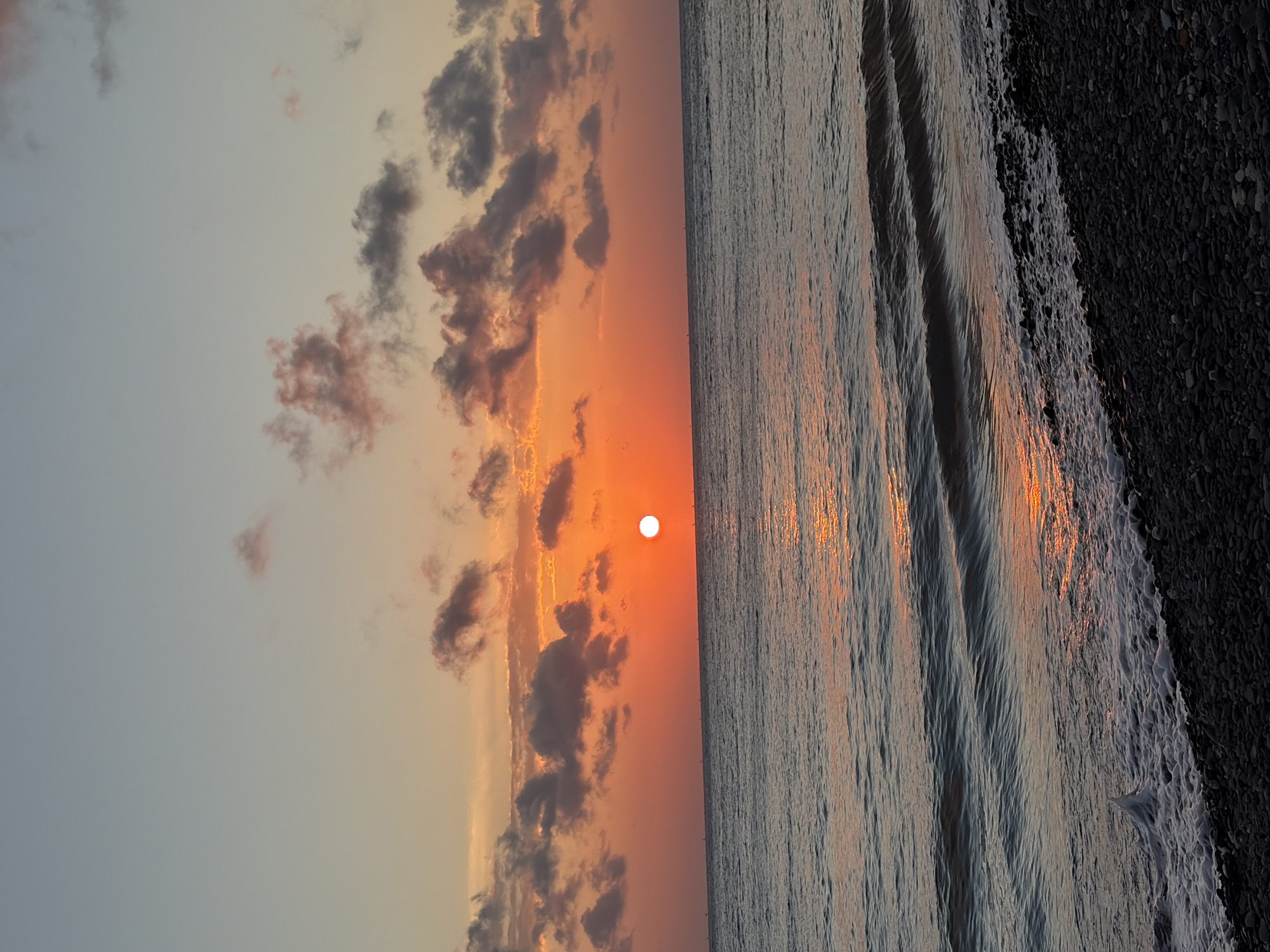 A serene sunset over the ocean is captured in this image, with the sun a bright, glowing orb just above the horizon. The sky transitions from a soft blue at the top to vibrant oranges and reds near the horizon, illuminated by the setting sun. Wispy clouds, tinged with pink and gray, are scattered across the sky, adding depth and texture to the scene. The ocean, a deep blue-gray, reflects the warm hues of the sunset, creating shimmering streaks of light on its surface. Gentle waves roll towards the shore, their white foam kissing the pebbled beach. The beach itself is covered in dark, smooth stones, creating a stark contrast with the vibrant colors of the sky and sea. The overall impression is one of tranquility and natural beauty. The image does not depict the city of Abang, Indonesia.