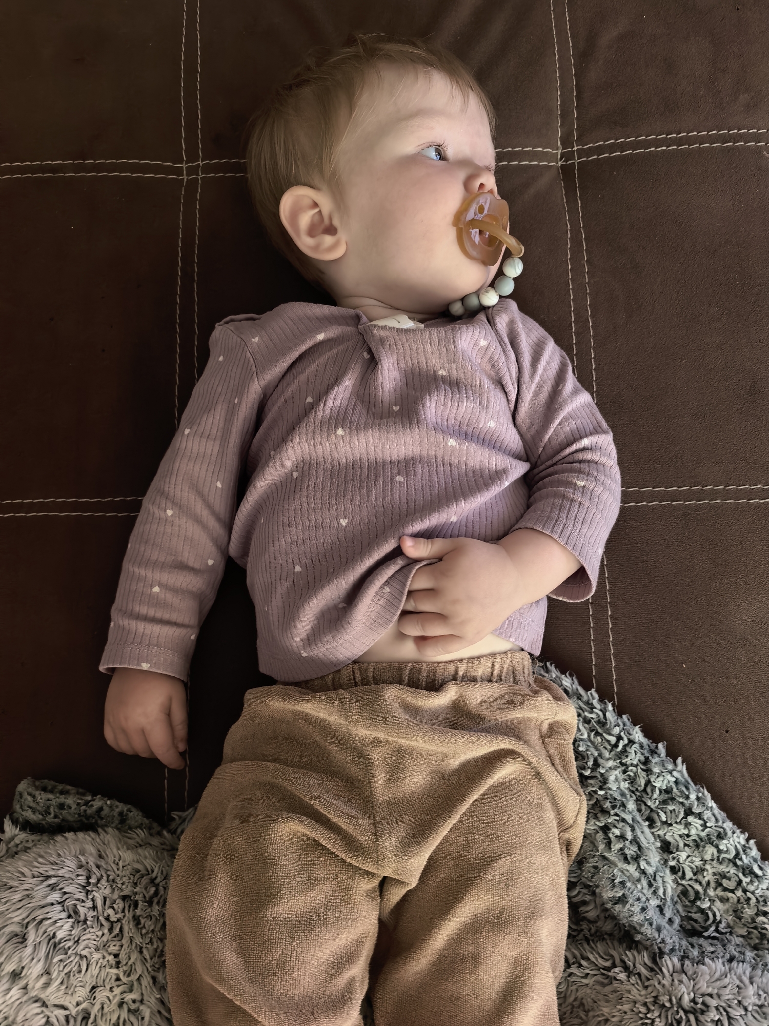 A baby is lying on a brown couch, looking to the side with a pacifier in their mouth. The baby is wearing a purple long-sleeved shirt with small white hearts and tan pants. A gray fuzzy blanket is partially visible on the right side of the image. The couch has visible stitching creating a grid pattern. The location of the image in Sterlitamak, Russia is unknown.