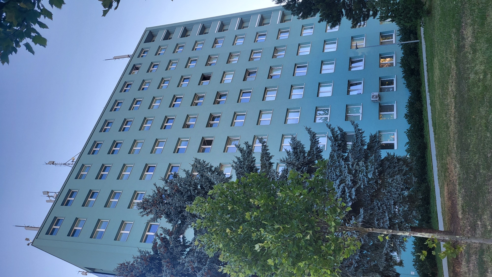 The image shows a multi-story residential building in Prešov, Slovakia. The building is painted a light teal color and has numerous windows with white frames. Several trees, including evergreen and deciduous varieties, are situated in front of the building. The sky above is clear and blue. Antennas and other communication equipment are visible on the roof of the building. The ground in front of the building is covered with grass and a paved walkway. There are no people visible in the image.