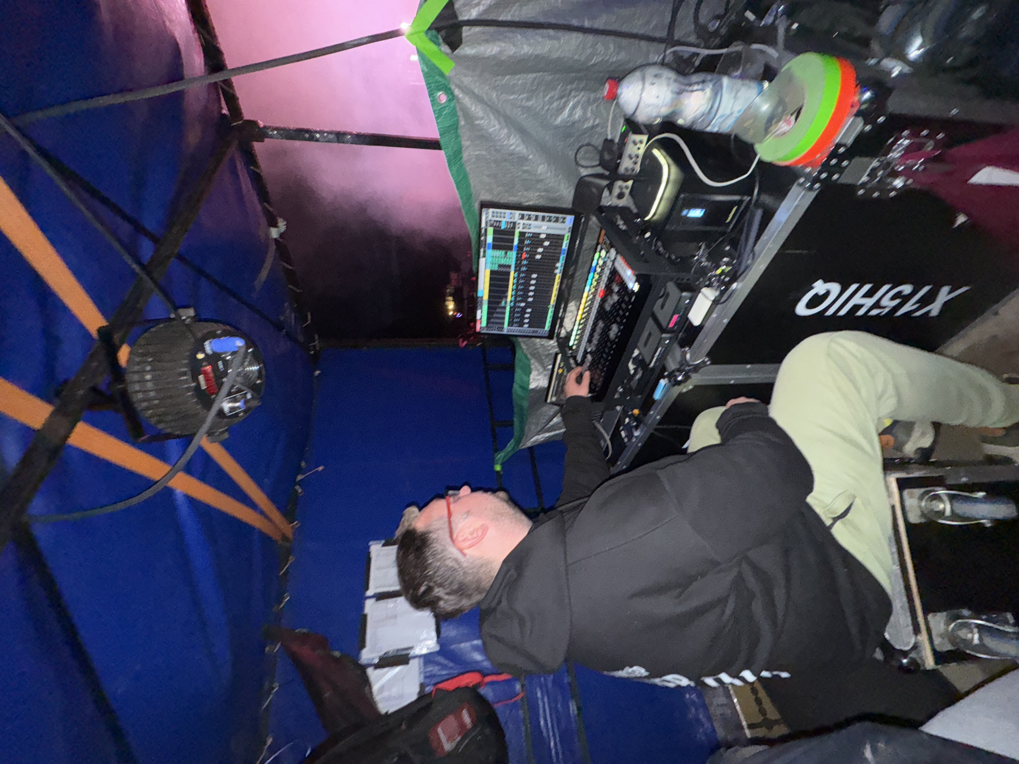 An image depicts an indoor, temporary event control booth at night. The image itself does not provide specific visual information to confirm the location as Bol'shaya Kivara, Russia.
A fair-skinned male individual, with short dark hair and wearing red-framed glasses, is seated on a black road case with wheels. He wears a black hooded sweatshirt and light green or khaki pants. His right hand rests on an external keyboard, positioned in front of an open laptop displaying what appears to be a digital audio workstation (DAW) or lighting control software with multiple faders and track elements.
The workstation setup includes several electronic devices, control surfaces, an external keyboard with multi-colored keys, and other black modular equipment. These items rest on a large black road case, which serves as a table, displaying "X15HIQ" in white lettering on its visible side. Cables connect the various components. To the right of the workstation, a clear plastic water bottle with a red cap is placed next to a black cylindrical object adorned with prominent green and red rings.
The booth's ceiling is constructed from a blue tarp, secured by crisscrossing orange straps, from which a reflective, faceted light fixture hangs. The left wall of the booth is also a blue tarp, with several white rectangular papers or notices taped to it, and a dark bag hangs nearby. The right side of the booth is enclosed by a gray tarp, which features a green strap at its upper right edge. Beyond the right side of the booth, an outdoor area is visible, brightly illuminated by a strong pink light, with atmospheric haze or smoke present.