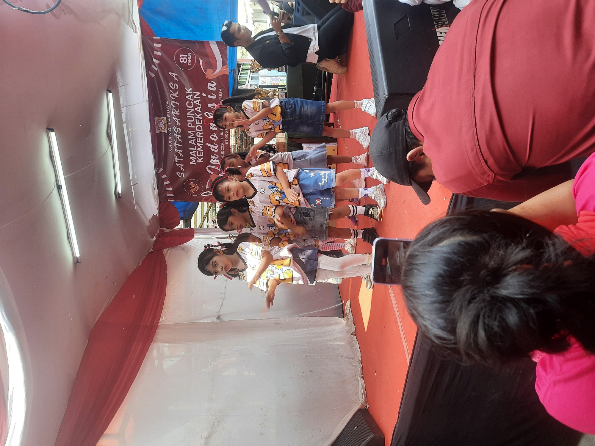 A group of young Indonesian girls are performing on a stage in what appears to be a temporary outdoor setup, likely for a community event. The stage is covered with a red carpet and is draped with red and white fabric, common colors in Indonesian flags, suggesting a patriotic celebration, possibly related to Indonesian Independence Day. A banner in the background reads "KARANG TARUNA SATATAS ARIKSA MALAM PUNCAK KEMERDEKAAN Indonesia" which translates to "Youth Organization Satatas Ariksa Independence Peak Night Indonesia." The number "81 TAHUN" (81 years) is also visible on the banner, likely referring to the anniversary of Indonesian independence.
The girls are dressed in matching t-shirts with cartoon characters and denim skirts or shorts, some wearing white tights. They are facing an audience, with one girl in the center actively gesturing with her hands. Several audience members are visible in the foreground, one of whom is holding up a smartphone to record the performance. To the right of the stage, a man in a black jacket is kneeling and looking at his phone, possibly a member of the event staff or an attendee. The environment suggests a sunny day, with ample lighting from overhead fixtures. The location is identified as Bandung City, Indonesia.