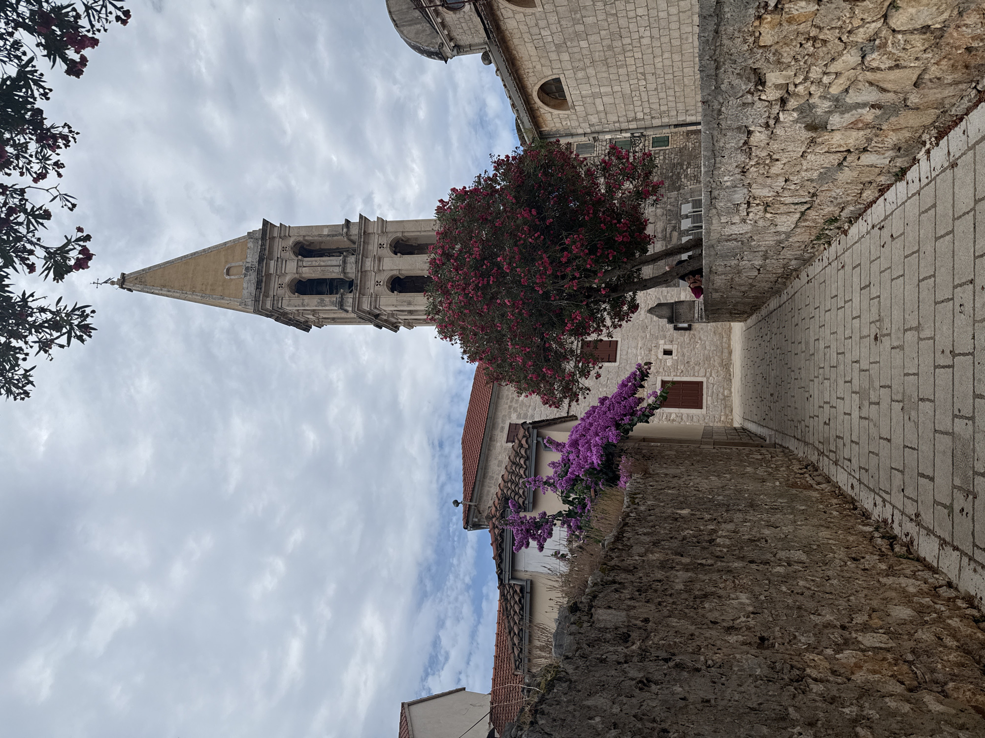 The image depicts a cobblestone pathway leading towards a historic stone church bell tower with a pointed yellow roof. The pathway is bordered by rough-hewn stone walls on either side. A vibrant tree with bright pink flowers stands to the left of the bell tower, partially obscuring the church's stone facade. Purple flowers cascade from a vine on the left side of the pathway, near the stone wall. The sky is overcast with dramatic clouds. The scene is located in the city of Grad Stari Grad, Croatia, as indicated by the architectural style and landscape typical of the region.