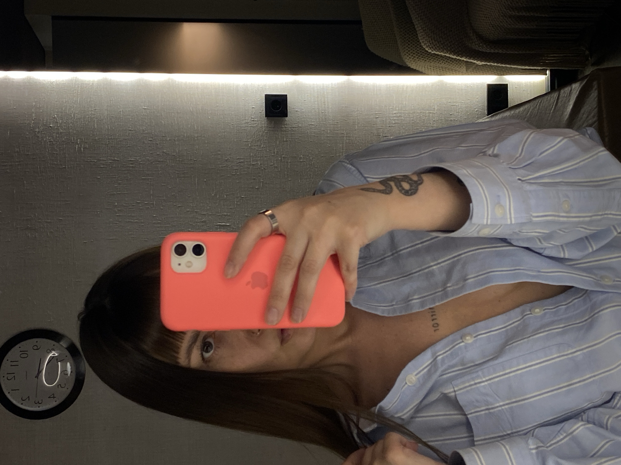 A woman is taking a selfie in a room with a clock on the wall. She is holding an orange phone in front of her face, and she is wearing a light blue and white striped shirt. The environment includes a wall with a light strip, what appears to be an object mounted on the wall, and a couch behind her. No information suggests the location is in Votkinsk, Russia.