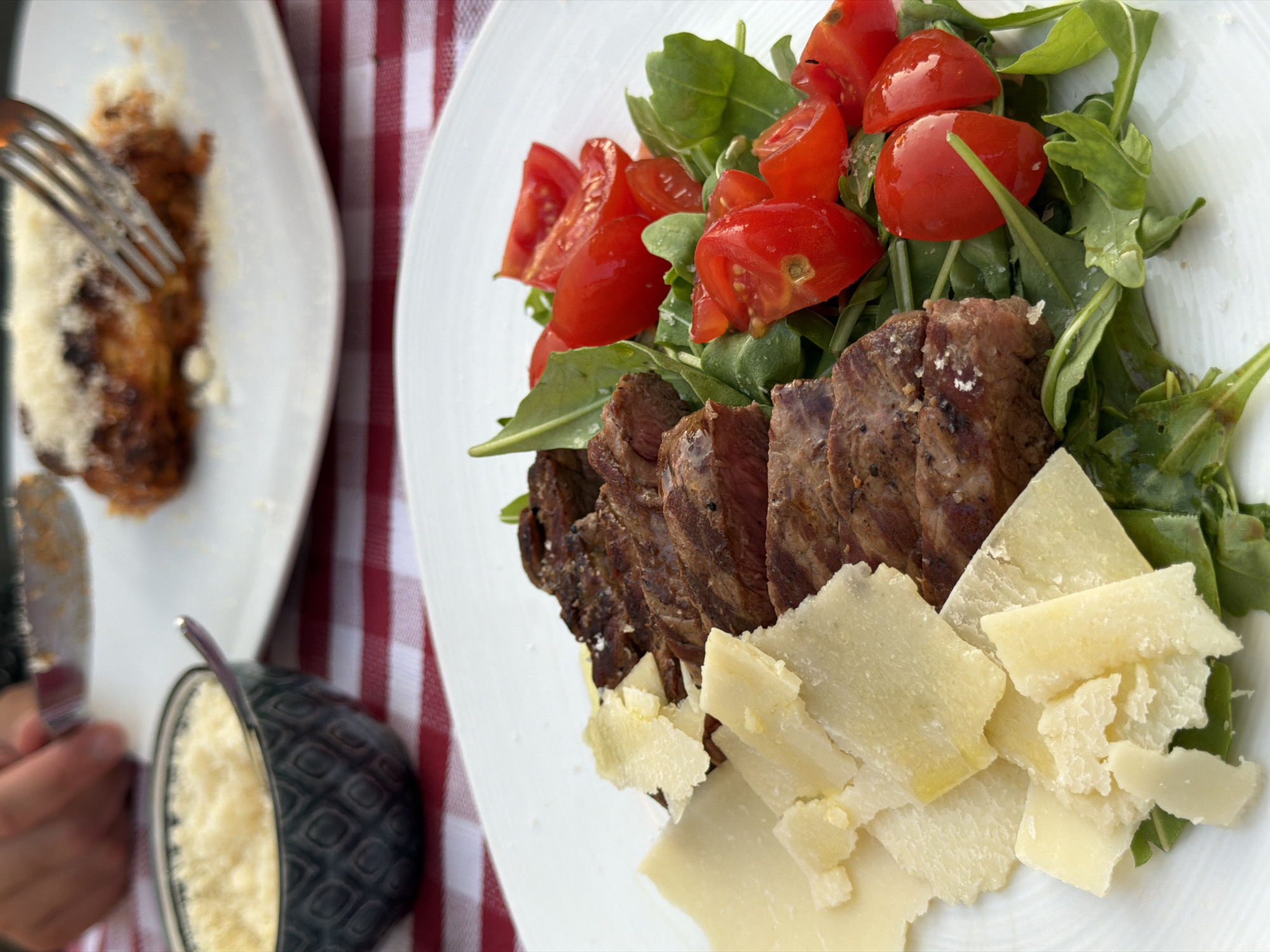 A close-up shot focuses on a plate of food served on a white tablecloth with a red and white checkered pattern. The plate contains slices of seared steak arranged next to a bed of arugula and cherry tomato halves. Shaved parmesan cheese is scattered over the steak and arugula, with larger shavings of parmesan cheese also placed to the left of the steak. In the background, another plate with what appears to be lasagna, topped with shredded cheese, is visible. A fork is partially inserted into the lasagna. A small dark bowl filled with shredded cheese and a spoon is positioned to the upper left of the frame, on the checkered tablecloth. The scene suggests a meal is being enjoyed at a restaurant in Sirmione, Italy, likely outdoors given the lighting.