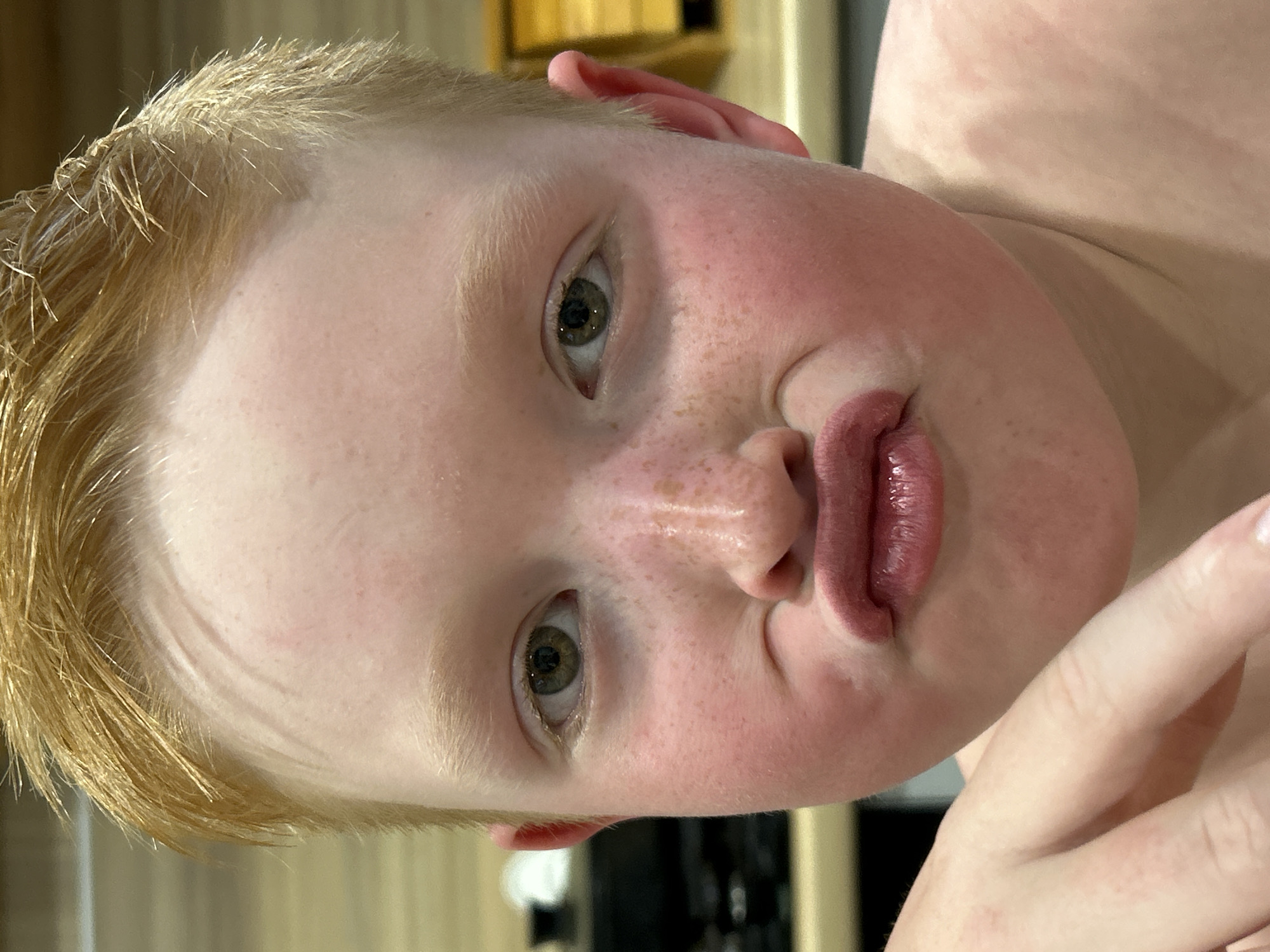 A close-up shot of a fair-skinned person with short blonde hair, green eyes, and a pouty expression. Freckles are visible on their face. Part of a hand is visible in the lower right corner of the frame. The location is unknown and is not indicated in the image.