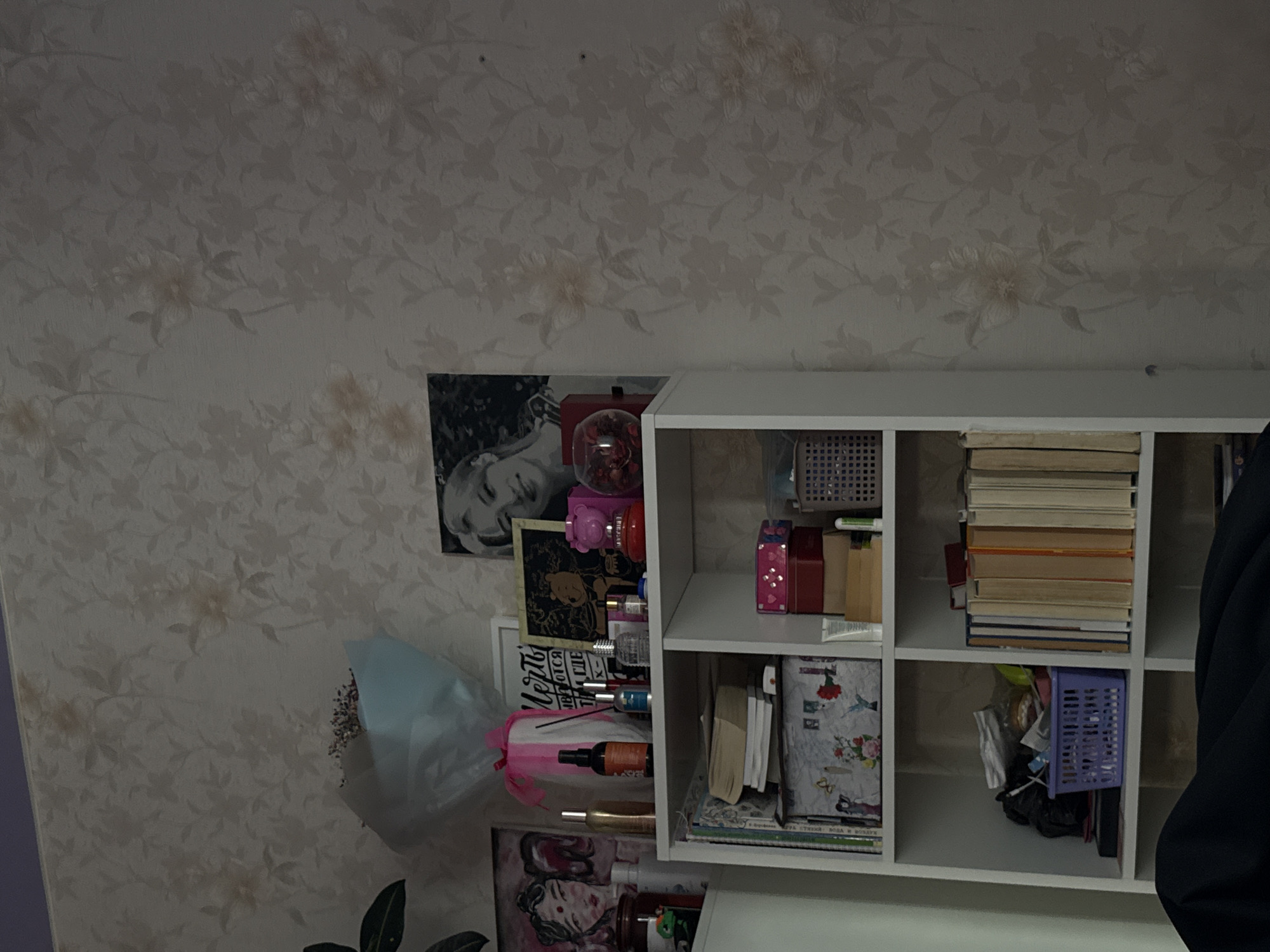 The image shows a section of a wall and a white shelving unit filled with various items. The wall is covered in floral wallpaper. Affixed to the wall next to the shelving unit are two portraits and a bouquet wrapped in light blue paper and secured with a pink ribbon.
The shelving unit contains books, storage baskets, cosmetic bottles, and other miscellaneous objects. A dark blue garment partially obscures the right side of the unit.
There's no explicit indication of the city of Perm, Russia, in the image.