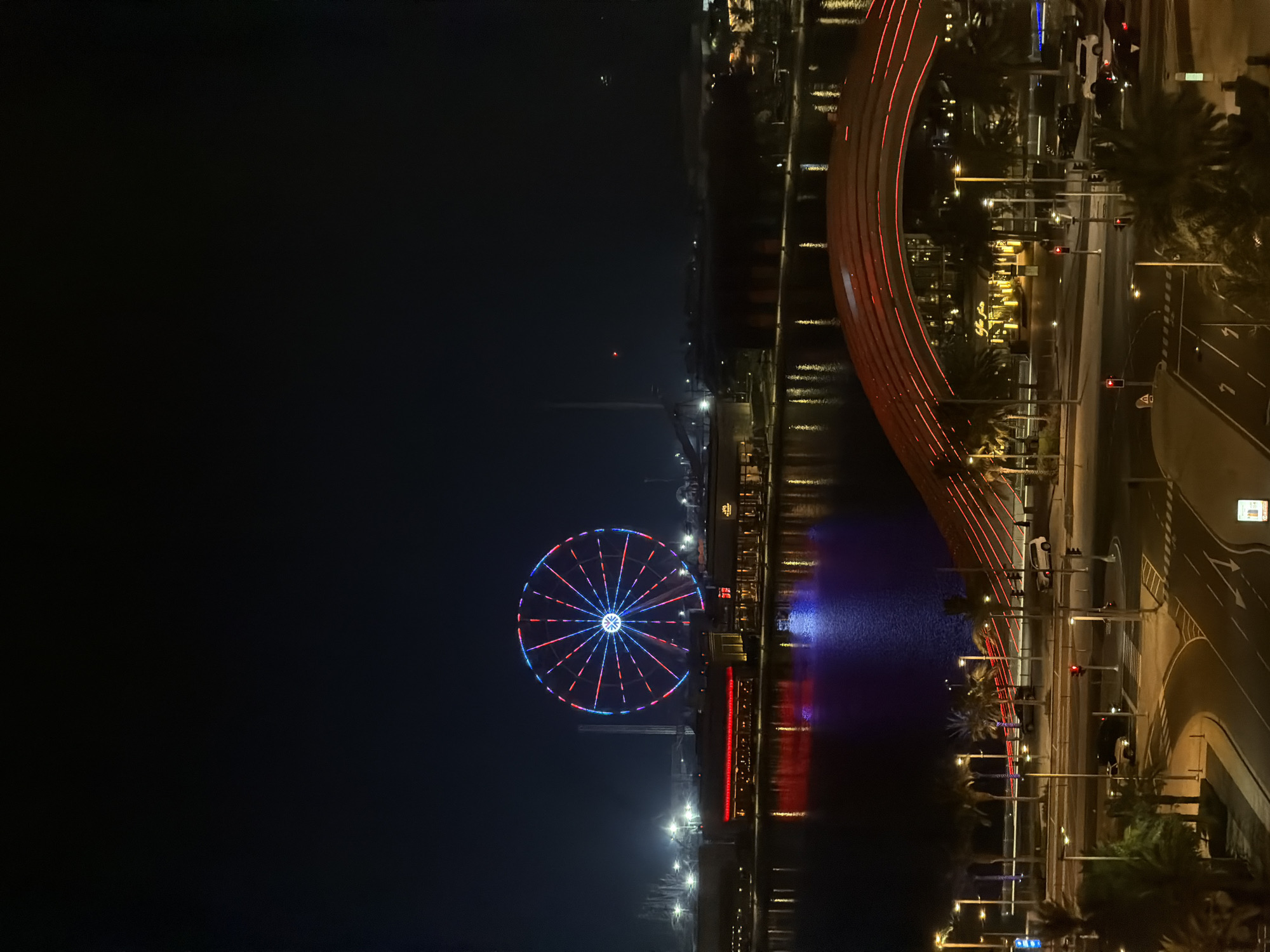 A nighttime, eye-level, medium shot captures an urban waterfront scene with a prominent Ferris wheel and a distinct bridge. The Ferris wheel, illuminated with blue, red, and white lights, is centrally located against a dark sky. In front of it, a body of water reflects the colorful lights of buildings lining the shore, which are also lit with red and blue hues. A modern, arching bridge, outlined in red lights, spans across the foreground, leading towards the water and buildings. Palm trees are visible along the waterfront and in the mid-ground. Below the bridge, a street scene unfolds with cars, streetlights, and crosswalks, indicating a busy urban environment. The overall impression is a vibrant and lively city at night, characterized by dynamic illumination and architectural features.
