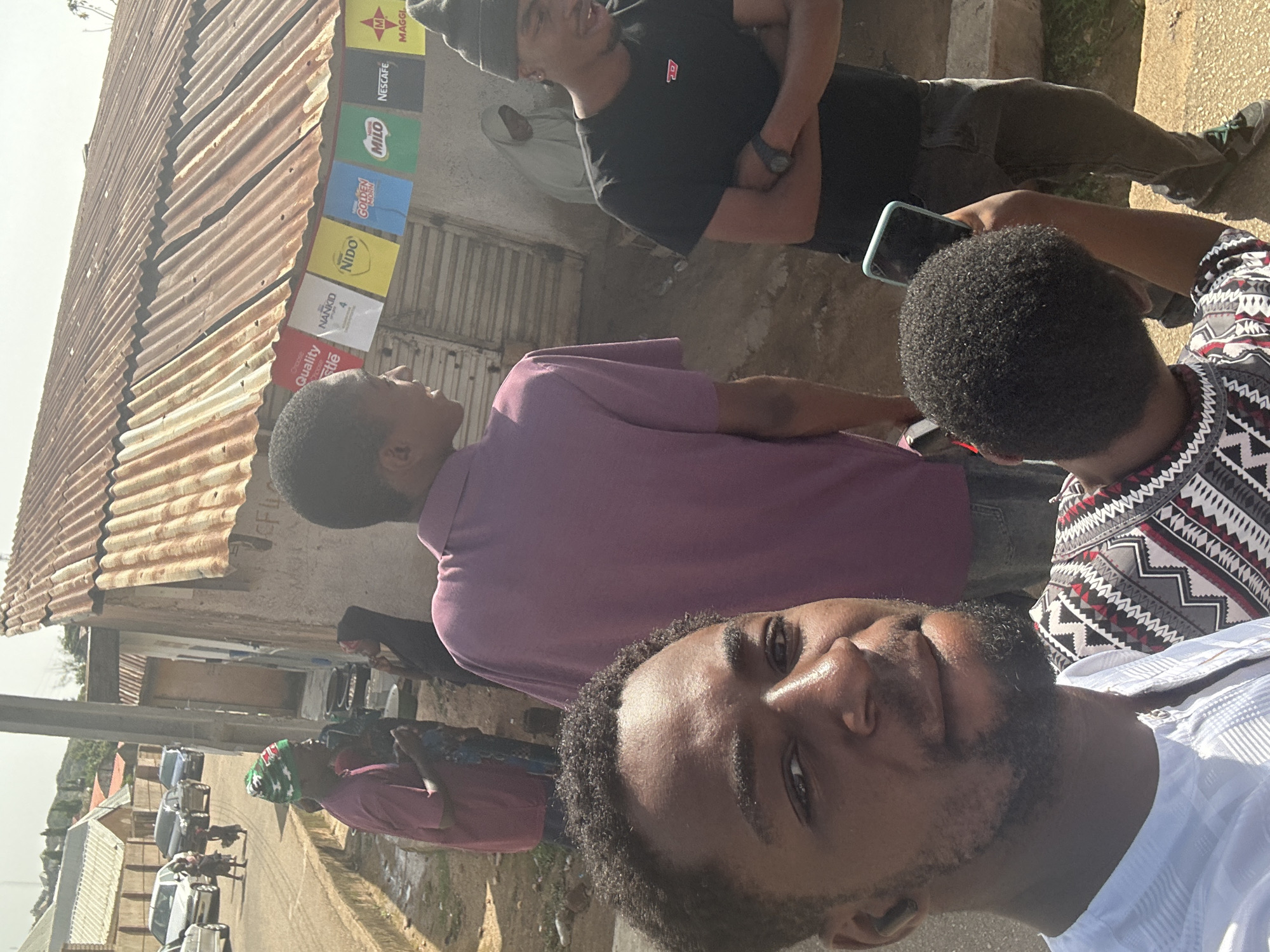 The image shows a group of men outdoors in an urban area. One man is taking a selfie, while others stand behind him. There is a building with a corrugated metal roof and advertisements for various products, including Maggi, Nescafe, Milo, Golden Morn, Nido, and others. A street with parked cars is visible in the background.