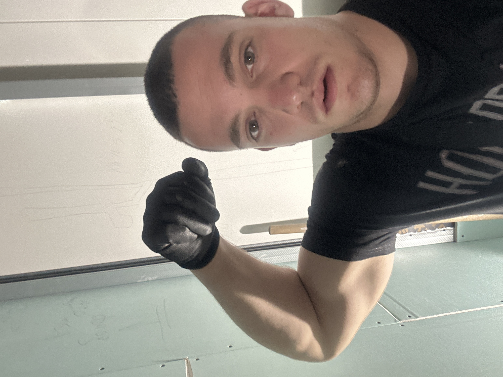 In a dimly lit interior space, a young man with a buzz cut and a black t-shirt poses for a selfie. He is wearing black gloves and flexing his right bicep. His left arm is out of frame. The background consists of light-colored walls, one of which appears to be drywall with some faint markings or writing. A metal support beam is visible to the right of the man. The scene suggests a construction or renovation environment. There is no visible indication of the location being in Burgas, Bulgaria.