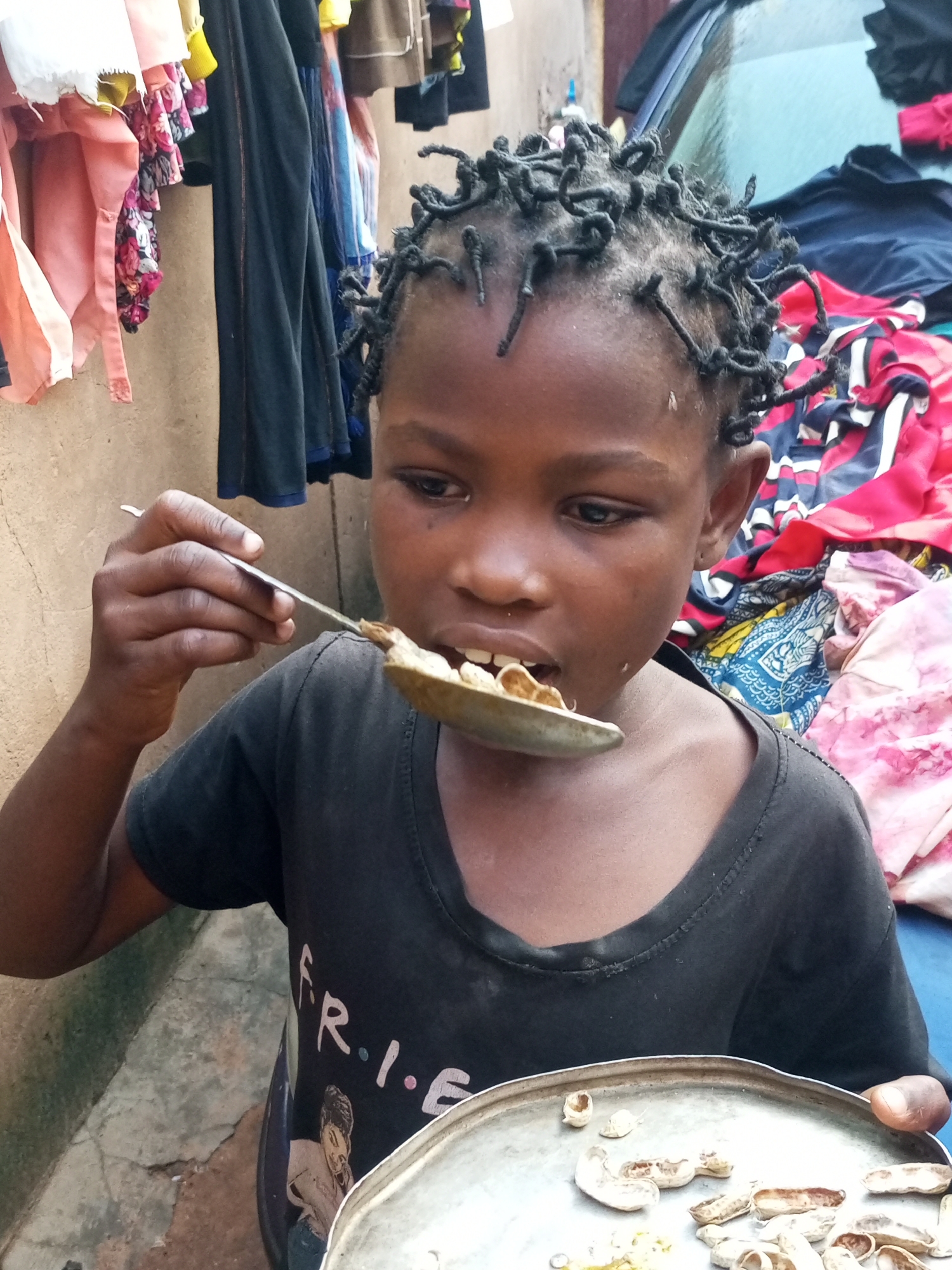 A young child is pictured outdoors in what appears to be Tempure, Nigeria, eating from a spoon and a metal plate. The child has dark skin, short, tightly braided black hair, and is wearing a black t-shirt with the word "FRIENDS" and an image of a person printed on it. The child is holding a metal spoon filled with food to their mouth. In front of the child is a metal plate containing several unpeeled peanuts and some shells. The background of the image shows a textured, light-colored wall and what appears to be laundry hanging or piled up, with colorful fabrics visible. The ground is concrete with cracks.