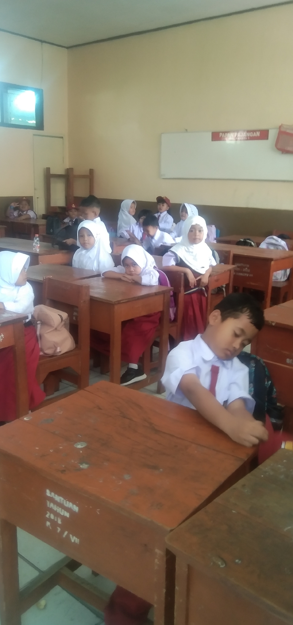 In a classroom setting, a group of young students, predominantly female, are seated at wooden desks. They are dressed in school uniforms consisting of white shirts and maroon skirts or pants, with many of the girls wearing white hijabs. The classroom appears to be brightly lit by natural light from a window on the left side of the image. Some students are looking towards the front of the room, while others are engaged in their work or resting their heads on their desks. The desks themselves are worn and appear to have some markings or writing on them, including text that reads "BANTUAN TAHUN 2018" and "R 7/VII". A whiteboard is visible on the wall at the front of the classroom, with a red sign that says "PAPAR PAJANGAN" (Display Board). The overall scene depicts a typical day in an Indonesian elementary school classroom. The specific location is not identifiable from the image alone, but the context suggests it is a public educational institution.