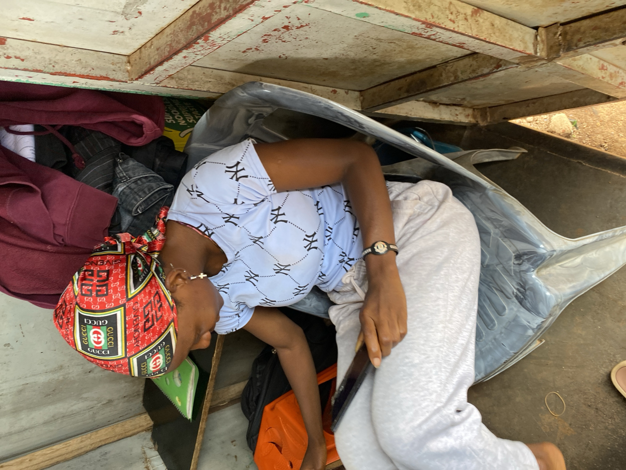 A person is partially visible within the confines of a confined space underneath a structure. The person is wearing a red headscarf with a "GUCCI" print, a light blue shirt with a pattern, and gray sweatpants. They are holding a dark-colored object in their hand, possibly a mobile phone. To the left of the person, there is a pile of clothing consisting of a maroon hoodie, black pants, and other garments. A green notebook or notepad is also visible. A large, gray object is positioned next to the person's legs. The structure above has wooden supports and a painted white surface. The location is Bukuru, Nigeria.