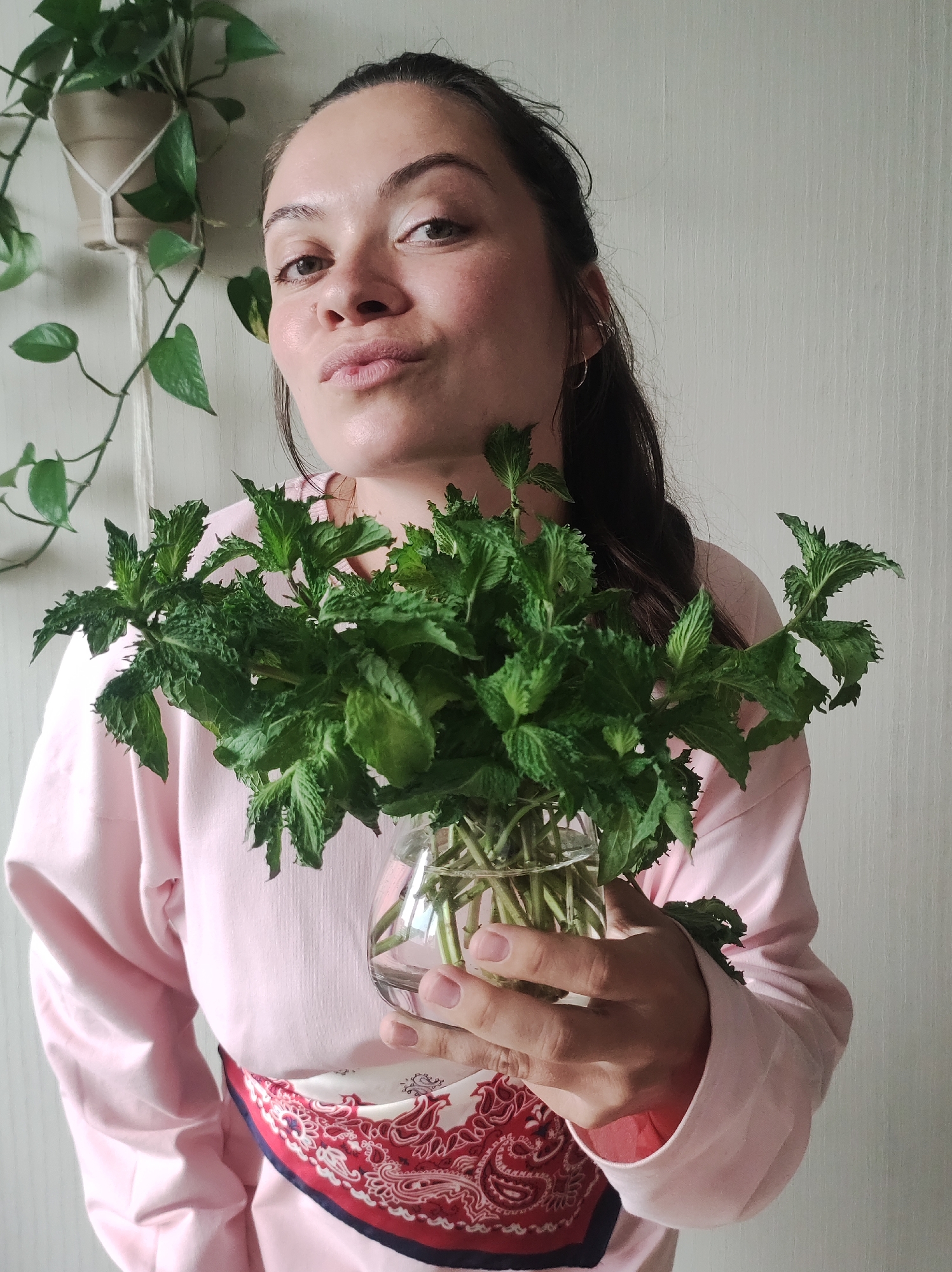 A young woman, possibly in her 20s, with dark hair pulled back, is holding a clear glass vase filled with fresh mint. She is wearing a pink sweatshirt and a red and white bandana is tucked into her waistband. She is looking at the camera with her lips pursed. Behind her, a potted plant hangs from a macrame hanger. The background is a textured, light-colored wall, suggestive of an indoor setting. The lighting is soft and natural. The scene appears to be in a residential setting, and could possibly be located in the city of Chaikovsky, Russia, given the context provided.