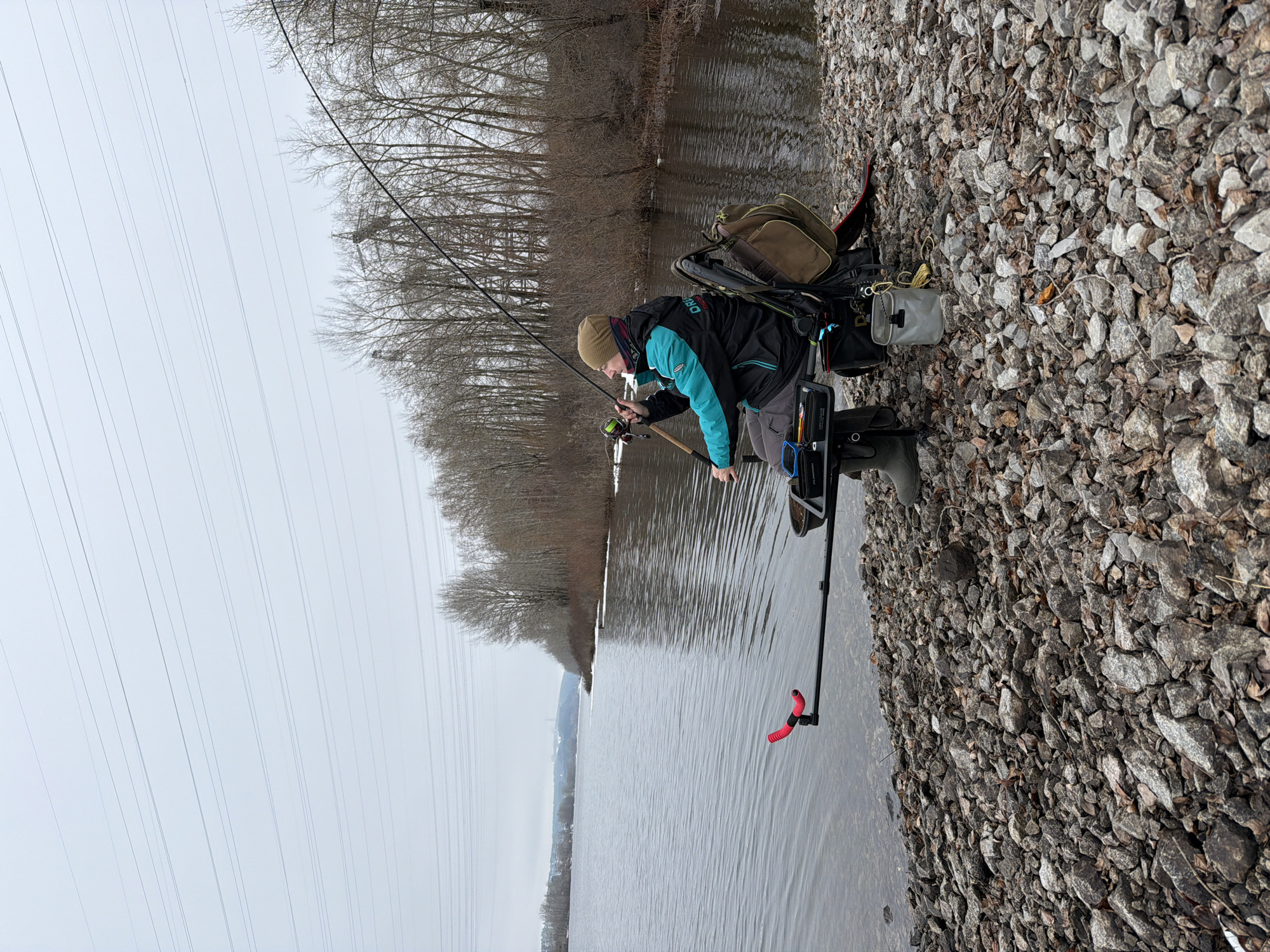 A man is fishing on the banks of the Kama River in Chaikovsky, Russia. He is wearing a dark jacket with light blue sleeves, grey pants, rubber boots, and a brown hat. He holds a fishing rod in his hands and is looking down at the water. To his right is a rocky bank. A fishing stand is in the water next to him. In the background, there are trees and electrical lines. The sky is overcast.