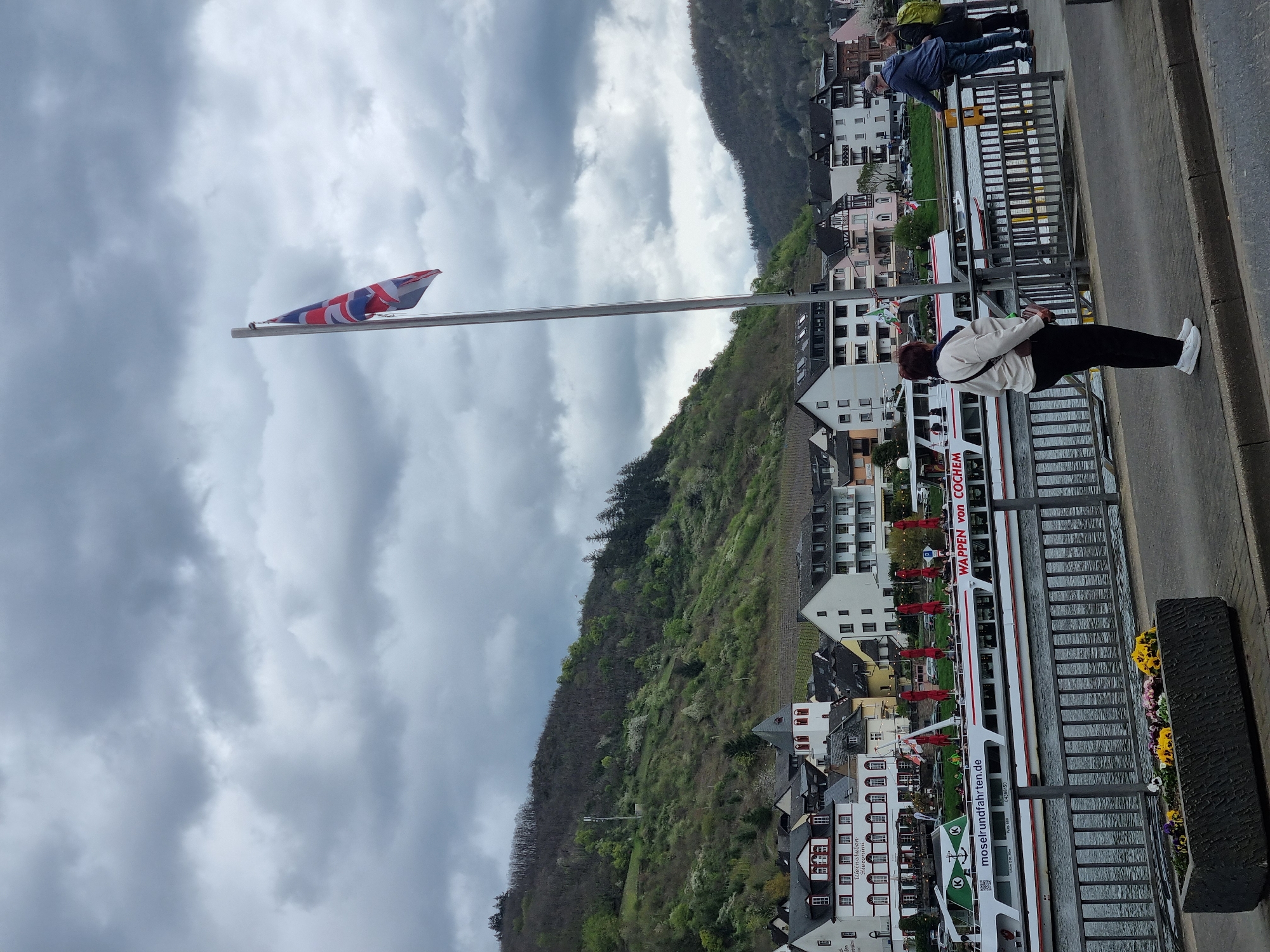 A vertical shot captures an outdoor scene in Cochem, Germany. A Union Jack flag is slightly unfurled atop a tall pole against a cloudy sky. Below, a hillside dotted with buildings and vegetation descends towards the river. Along the riverbank, a tourist boat, labeled "WAPPEN von COCHEM", is docked, with the text "moselrundfahrten.de" visible. Two people are on the right side of the image, leaning over a railing overlooking the river. One is wearing a white jacket and black pants, while the other person further away is wearing blue and gray clothes. A flower box rests on the edge of the sidewalk. The overall atmosphere is slightly overcast.