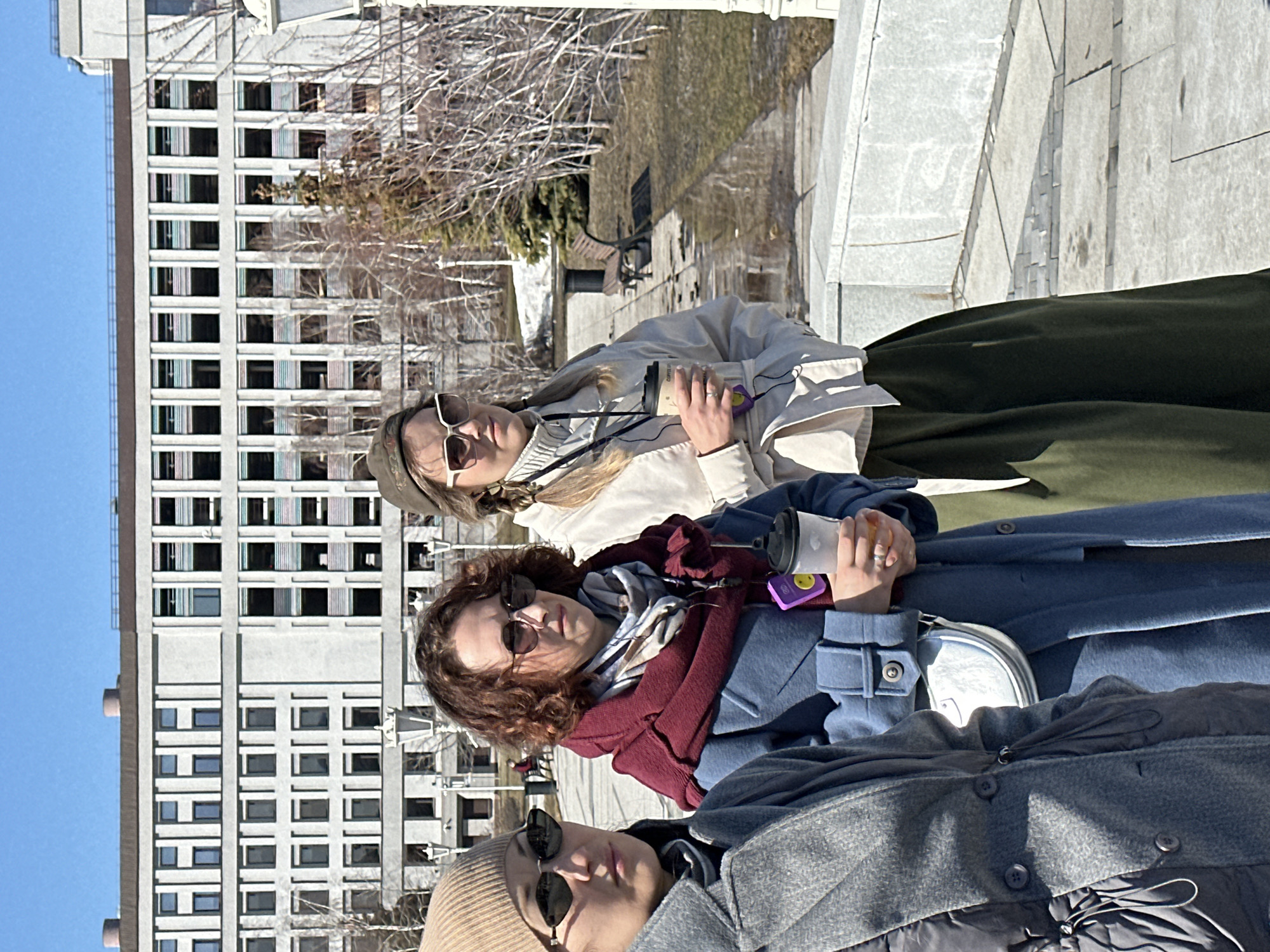 Three young women are posing outdoors, likely in Kazan, Russia, given the urban architectural style in the background. The woman on the top is wearing a light-colored jacket and a green skirt. The woman in the middle is wearing a blue coat with a red scarf and sunglasses. The woman on the bottom is wearing a gray jacket with a beige beanie and sunglasses. Each of them holds a coffee cup. The background features a multi-story building and bare trees. The setting suggests a sunny day in early spring.