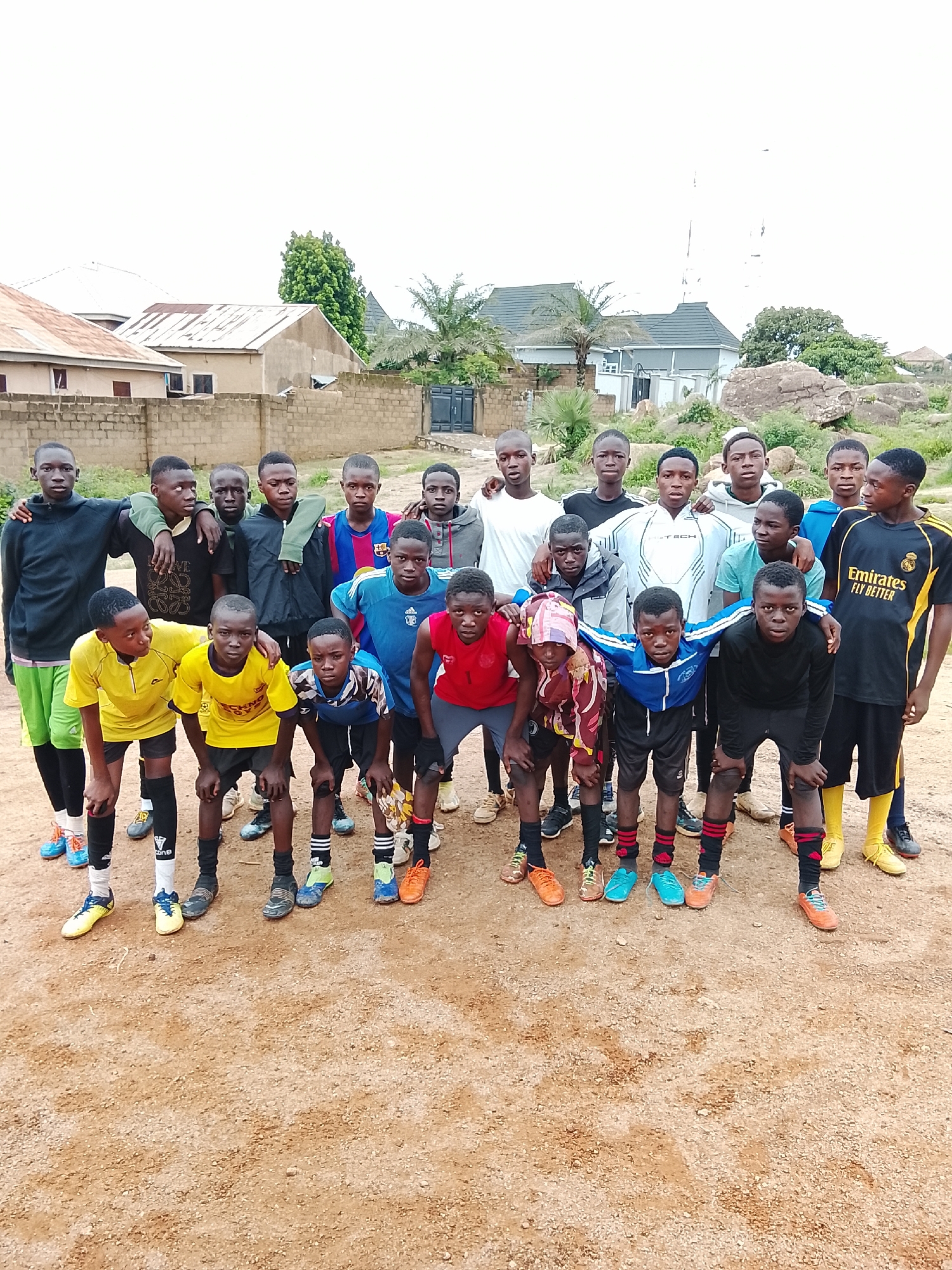 A group of 18 young males, appearing to be of African descent, are posing for a photograph outdoors. They are dressed in various athletic attire, primarily soccer uniforms, with some wearing t-shirts and athletic pants, and others in shorts. The majority of the group are wearing soccer cleats. The individuals are arranged in two rows, with the front row crouching and the back row standing. Some of the subjects have their arms around each other.
The background consists of a dirt or sand-covered ground. Behind the group, there is a brick wall and several buildings with corrugated metal roofs, suggesting a residential area. Further in the background, there are trees and rocky outcrops, indicative of a semi-arid or savanna landscape, consistent with the environment of Jos, Nigeria. The sky is overcast and bright. There are no obvious signs of immediate danger or distress. The overall scene depicts a gathering of young people, likely a soccer team, in an outdoor urban or semi-urban setting in Jos, Nigeria.