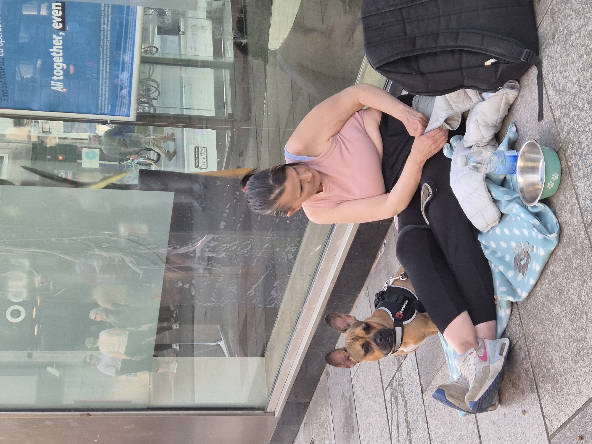 A medium shot from an elevated perspective captures a woman and her dog on a sidewalk in Cardiff, United Kingdom. The woman, with dark hair pulled back in a ponytail, is wearing a light pink tank top, a blue sports bra, and black leggings. She's sitting on the sidewalk, surrounded by a black backpack, a water bottle, a silver dog bowl with a green exterior, and a blue and white patterned blanket. Her arms are occupied as she seems to be adjusting or handling an object in her lap. On her feet are mismatched sneakers.
A brown dog wearing a black harness labeled "barkhaus" is seated beside her, looking directly at the camera with attentive eyes.
The setting is an urban environment, as suggested by a large glass window reflecting people walking by. An advertisement or public announcement is visible on the upper left corner of the window. The sidewalk appears to be made of rectangular stone tiles.