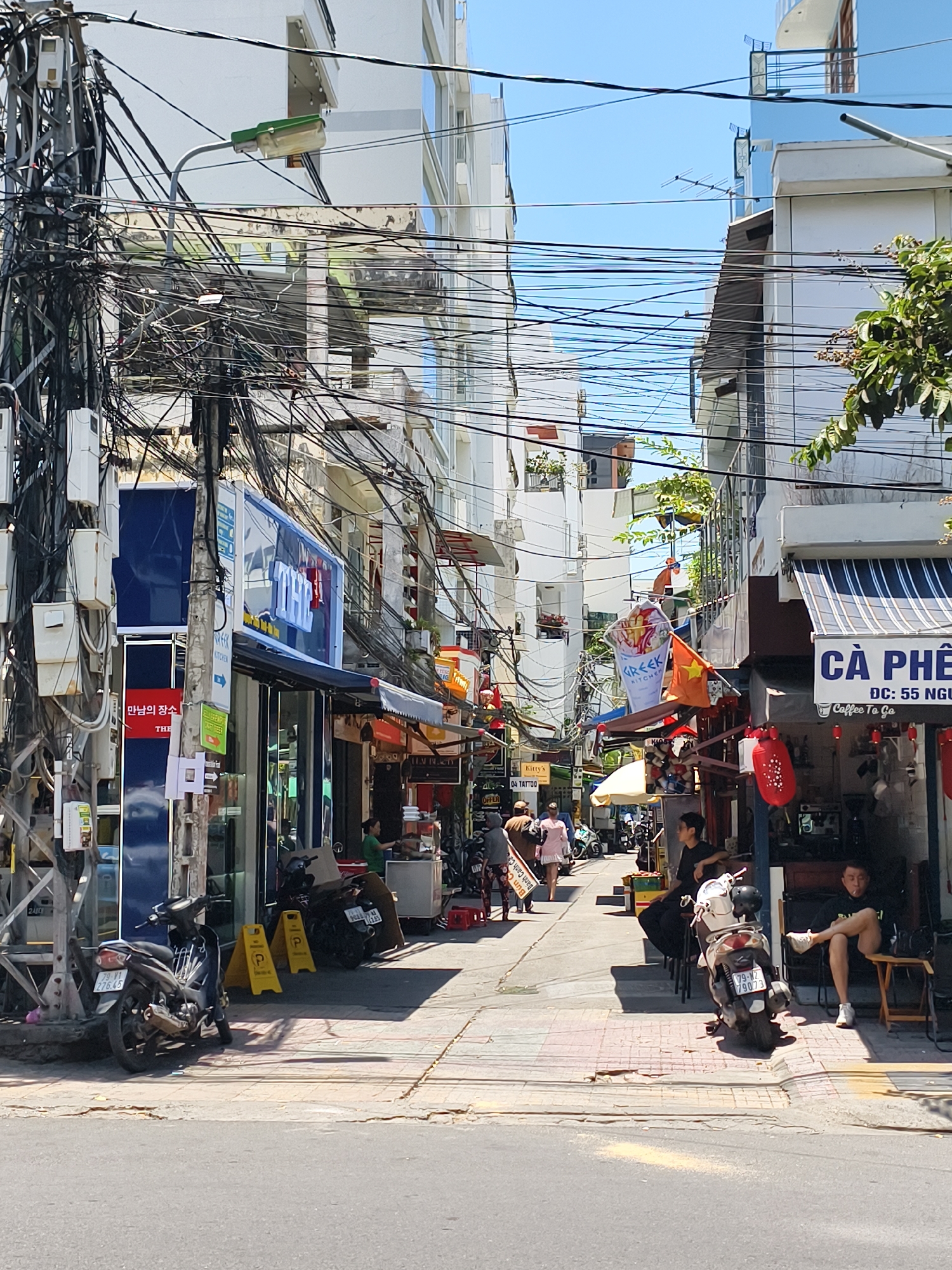 The image depicts a narrow street in Nha Trang Ward, Vietnam, characterized by dense overhead cables. The street is lined with storefronts, including a cafe labeled "Cà Phê" and a restaurant with the Korean script. Several people are walking along the street, and others are sitting outside the cafe. Motorbikes are parked along the sides of the street. A sign for "Kitty's 04 Tattoo" is visible. Banners, including one for "Greek Kitchen," and Vietnamese flags are displayed.