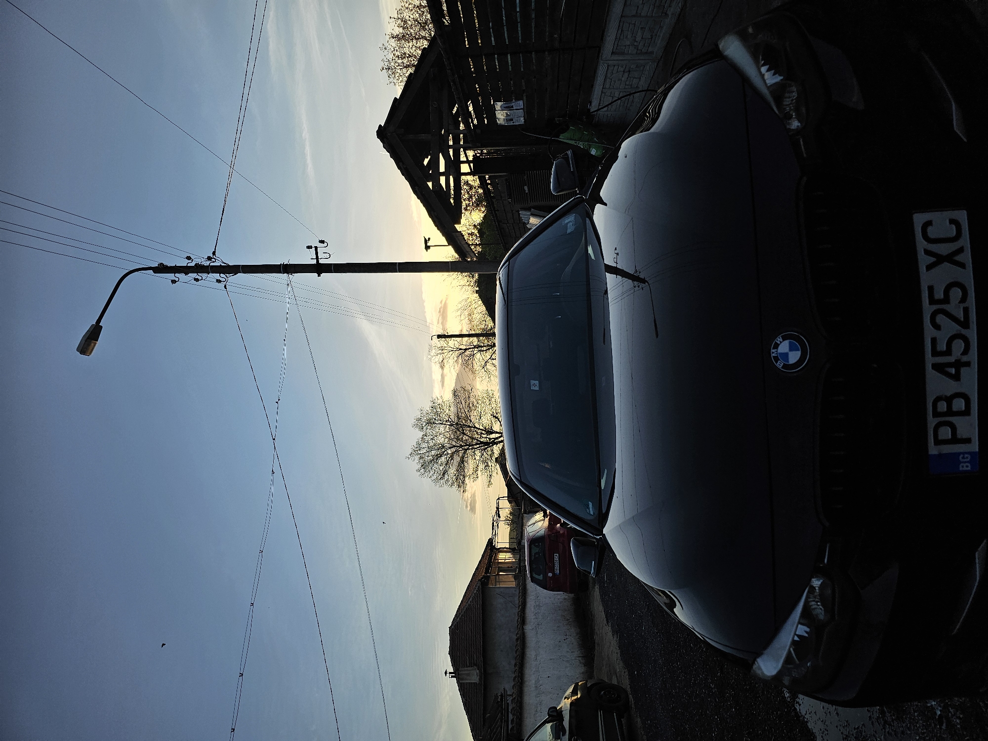 The image shows a black BMW car parked on a street in Krasnovo, Bulgaria. The license plate reads "PB 4525 XC". The car is facing the right side of the frame. The sky is a light blue gradient, suggesting it may be sunset or sunrise. A utility pole with a street lamp is visible on the left side of the frame. In the background, houses and trees are visible. A wooden structure with a roof is also present in the mid-ground. Other vehicles appear to be parked behind the BMW, including a red car.