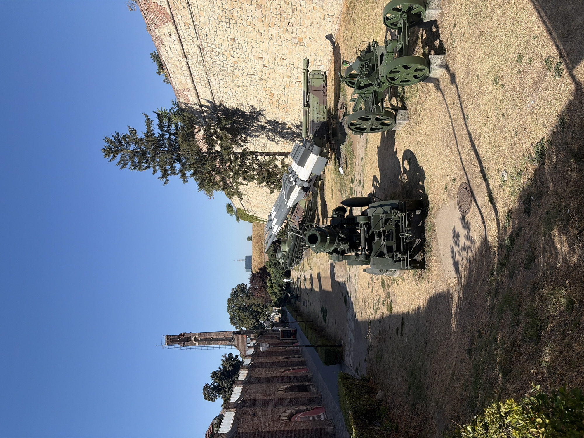The image depicts an outdoor display of military equipment within the Belgrade Fortress in Belgrade, Serbia. The foreground features several green artillery pieces, including a large mortar and a towed gun. In the middle ground, a missile launcher system is visible, along with a tank. The background is dominated by the fortress walls, constructed from stone and brick. To the left, a tall, brick clock tower stands against a clear blue sky. The ground is covered in dry grass and dirt, with scattered shadows indicating a sunny day. The scene appears to be a historical exhibit, showcasing weaponry from different eras. No people are clearly visible interacting with the exhibits.