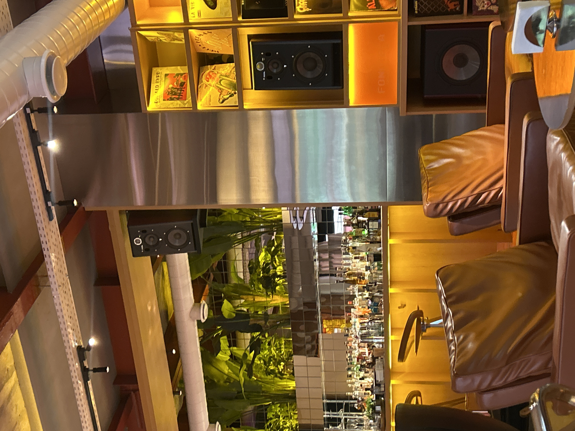 This media file captures an interior scene, likely a bar or lounge, characterized by a retro-modern aesthetic. No people are visible.
On the left side of the image, white corrugated ventilation ducting runs horizontally, with a black metal channel featuring small spotlights below it. The central focus is a large, illuminated shelf unit with various compartments. Some shelves display what appear to be vintage vinyl records or album covers, one of which clearly shows the text "DO AD BYPD". Other compartments house black audio speakers, including both bookshelf-sized monitors and a larger subwoofer. An orange rectangular panel within the shelving unit has partially visible text that appears to be "FONOS".
Below the main shelf unit, a reflective, possibly mirrored or polished metal surface extends horizontally. To the right, several comfortable-looking bar stools or chairs are arranged, featuring brown leather cushions and dark frames.
The lower left portion of the image reveals lush green plants, possibly in planters, adding a natural element to the decor. To the lower right, a well-stocked bar area is visible, featuring numerous bottles of alcohol arranged on shelves, reflecting in a mirrored back wall.
The scene is illuminated by warm, artificial lighting, suggesting an evening or nighttime setting. The combination of music-related decor (records, speakers), comfortable seating, and a bar points to a social establishment designed for relaxation and entertainment. The overall ambiance appears inviting and stylish. The location context provided is Yerevan, Armenia.