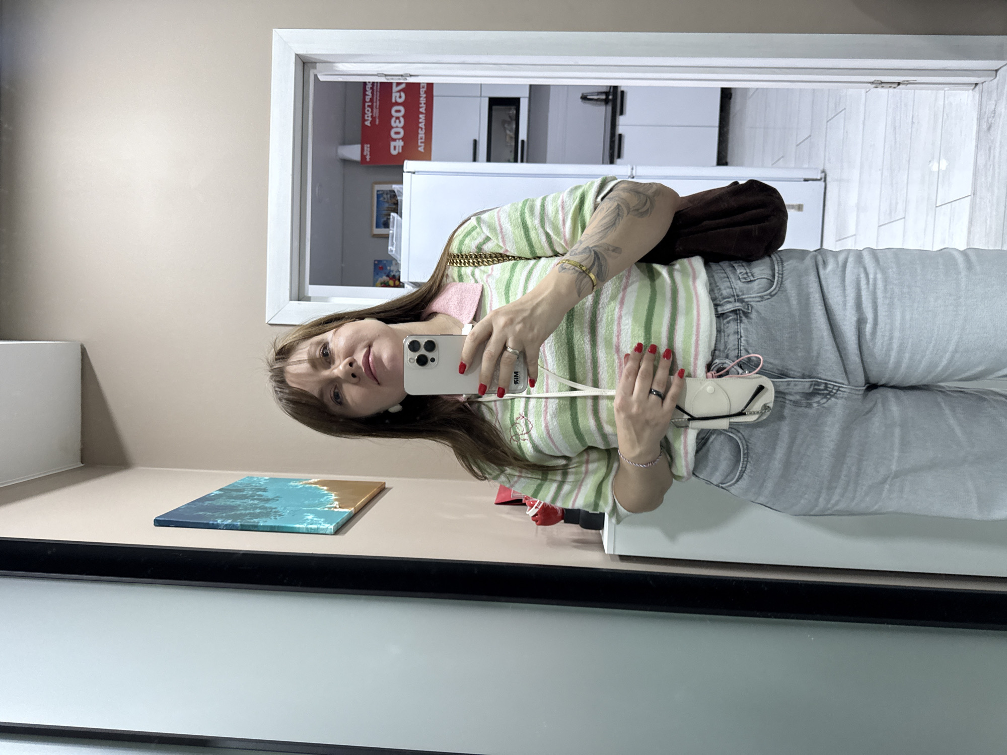 A medium-shot selfie of a Caucasian woman is taken in a well-lit, indoor environment that appears to be a home or apartment. The woman, who has long brown hair and is wearing a striped green and white long-sleeved shirt with a pink collar, is holding a white smartphone in her right hand, angled to capture her reflection. She is also wearing light blue denim jeans and a dark brown purse is slung over her left shoulder. Her nails are painted red, and she has several tattoos on her left arm. A white phone case with a pink string is attached to her jeans. In the background, a doorway reveals a modern kitchen area with white cabinets and appliances. On the wall to the left of the woman, a colorful abstract painting of a beach and ocean scene is visible. The overall setting suggests a casual, domestic scene. There is no identifiable context that places this image specifically in Sochi, Russia.