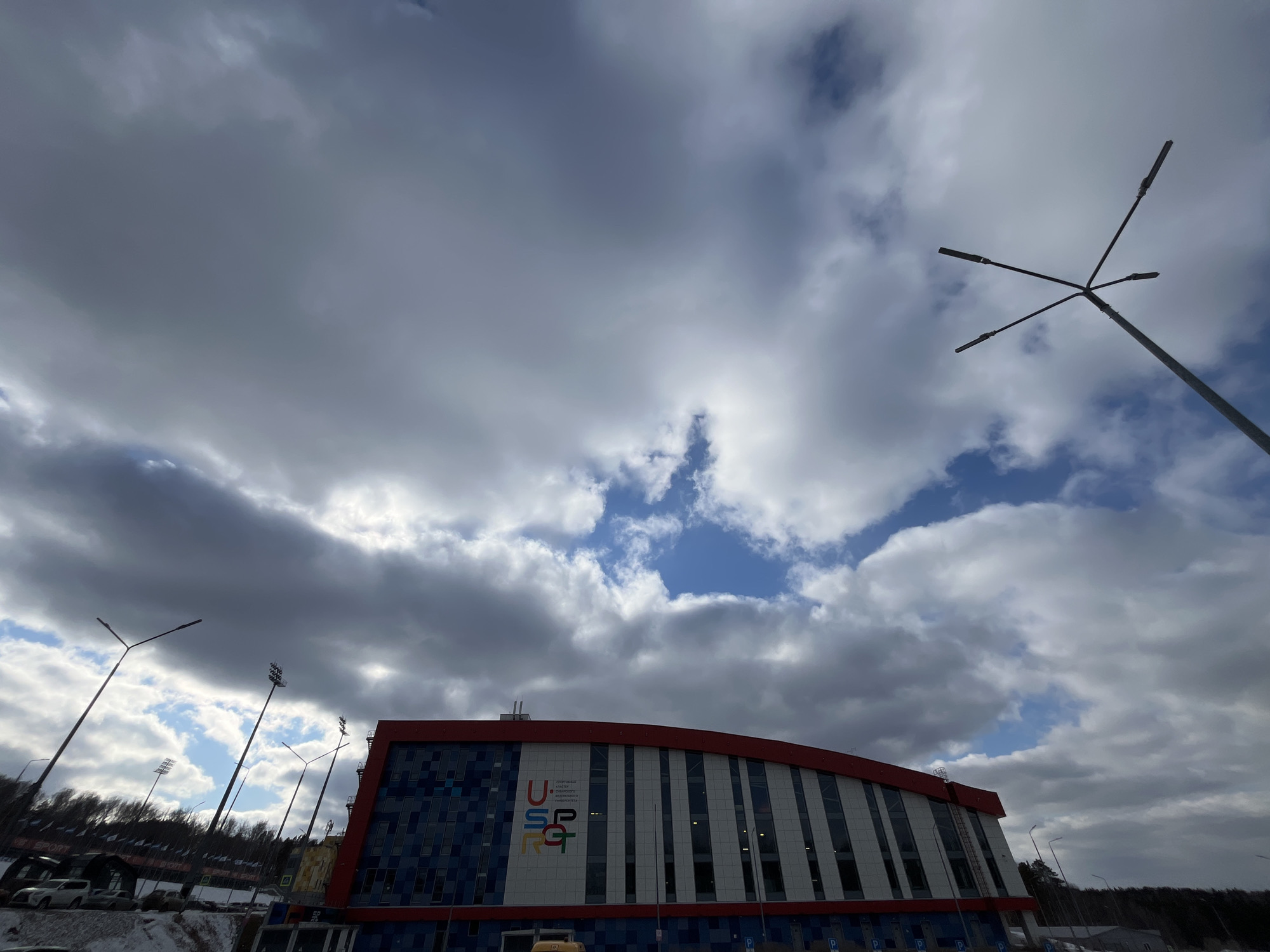 The image shows the Ice Arena "Pervomaysky" in Krasnoyarsk, Russia, on a cloudy day. The arena has a red roof, blue and white exterior walls, and the logo "U SP RGT" on its facade. Several light poles are visible around the building. The ground shows signs of snow cover, indicating a cold environment. In the background, a forest line can be seen. Cars are parked near the arena.