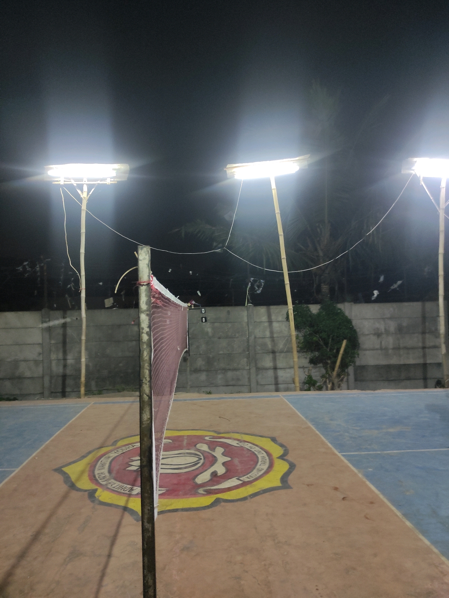 An outdoor badminton court is illuminated by bright lights at night. A net stands in the foreground, dividing the court. The court surface is marked with different colors and a circular emblem. A concrete wall forms the background, with trees and foliage visible behind it. The location is identified as Padalarang, Indonesia.