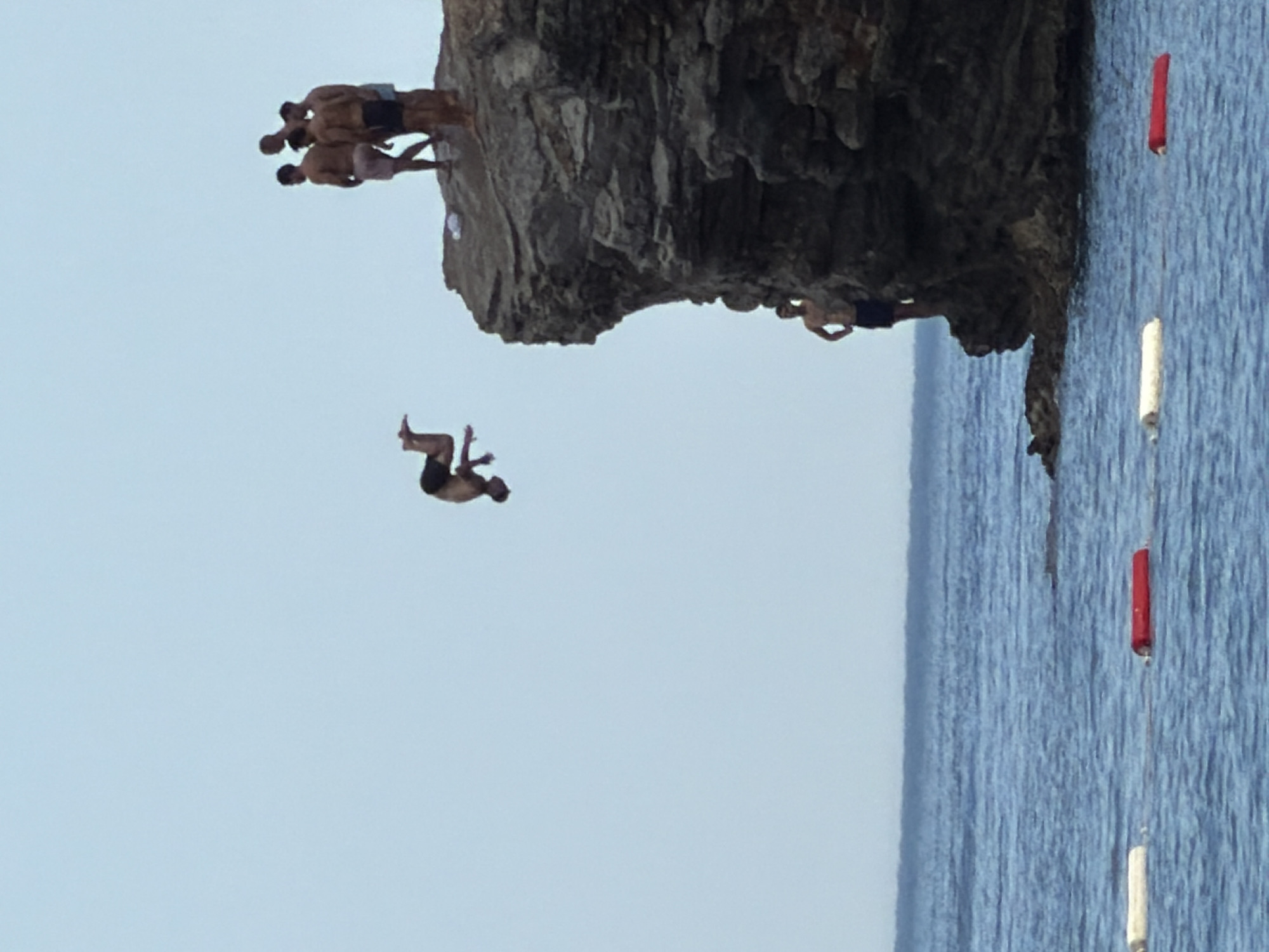 The image depicts a clear day with a light blue sky and calm blue water, likely the Adriatic Sea. In the foreground, several buoys, one red and one white, float on the water's surface. The main focus is on a rocky cliff face where a group of young men are engaged in cliff diving. One individual is mid-air, performing a somersault, while several others stand at the edge of the cliff, observing or preparing to jump. Another young man is positioned on a lower ledge of the cliff, looking out at the water. The scene appears to be located in Budva, Montenegro, a popular destination known for its stunning coastline and opportunities for adventurous activities. The overall atmosphere is one of youthful energy and daring.