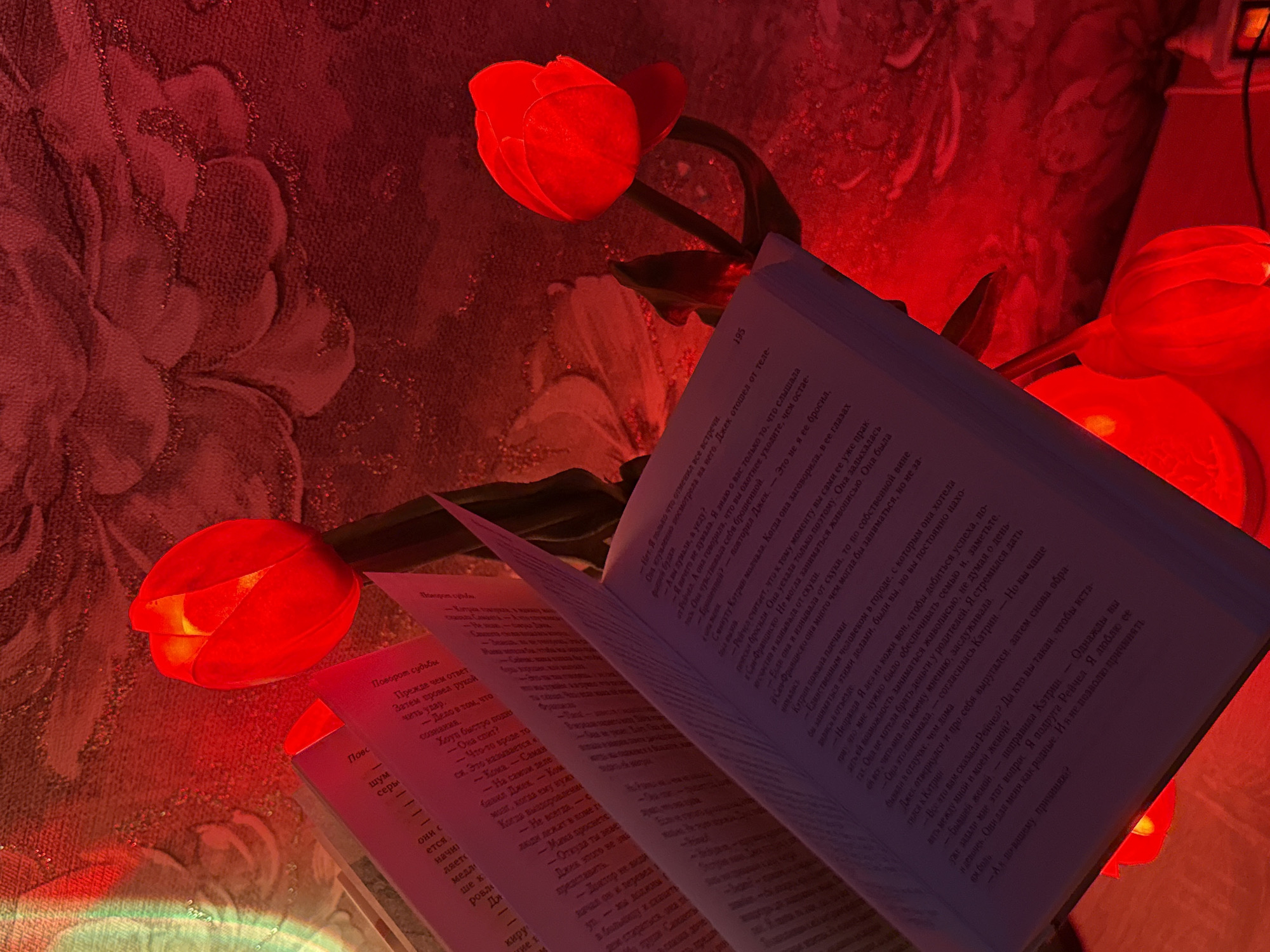 An open book is illuminated by red light from artificial tulips. The book is positioned on a textured, possibly fabric, surface that appears to be a wall or tapestry, with a floral pattern visible through the red lighting. The tulips are glowing from within, casting a strong red hue over the scene. The image is a close-up shot, focusing on the interplay of light and shadow on the book and surrounding elements. There are no people or identifiable locations present.