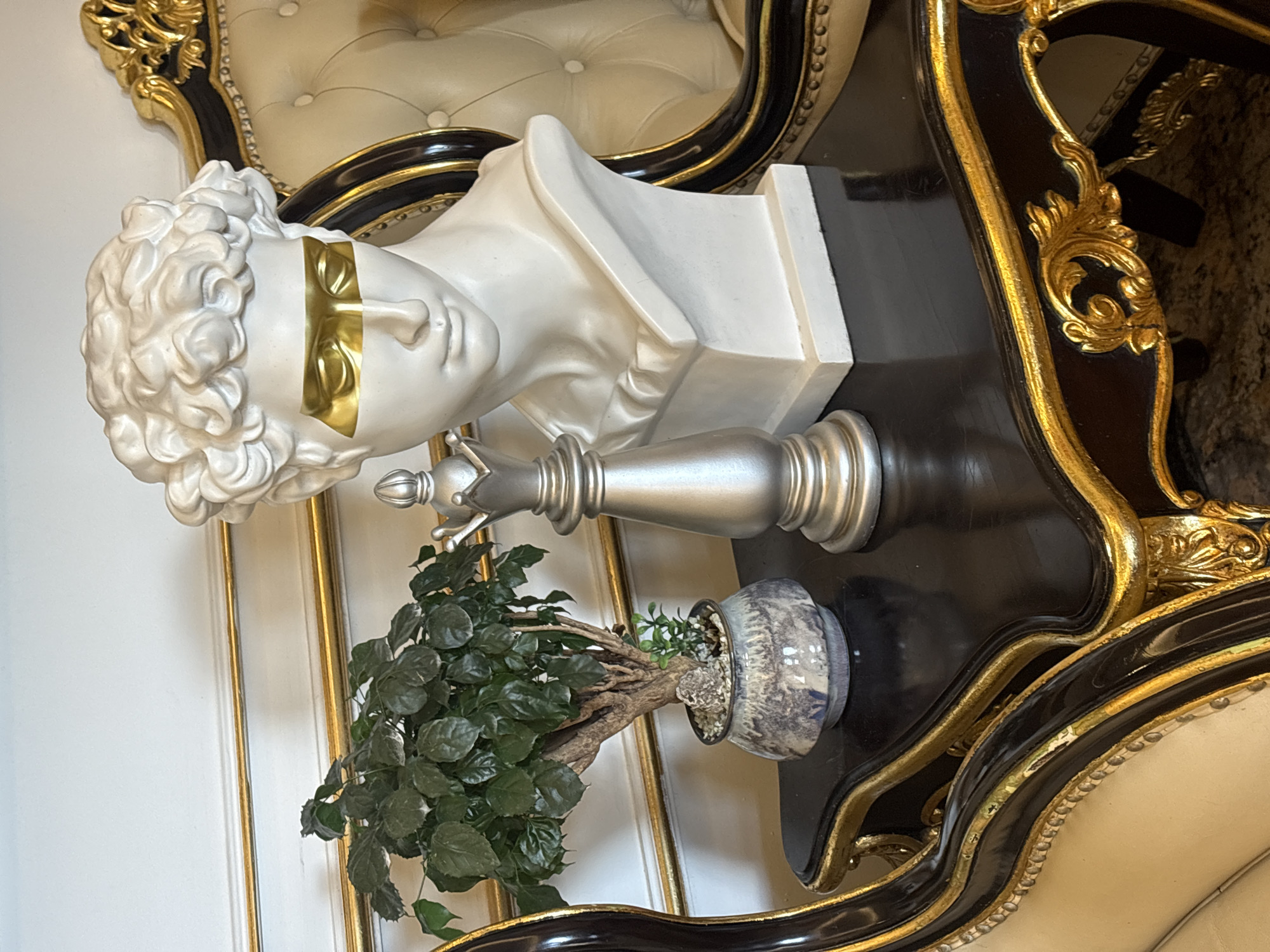 The image depicts a still life composition on a dark, ornate table with gold accents. The table is placed in an indoor setting, possibly a room with white walls adorned with gold trim. On the table are several decorative objects: a white bust of a classical figure with gold bands across its eyes, a silver chess piece resembling a queen, and a small potted plant with lush green leaves. The background includes a glimpse of a tufted cream-colored upholstered chair with gold detailing, suggesting a luxurious interior. The scene is well-lit, highlighting the textures and finishes of the objects. There is no indication of Đà Nẵng, Vietnam in the image; it is a generic indoor scene.